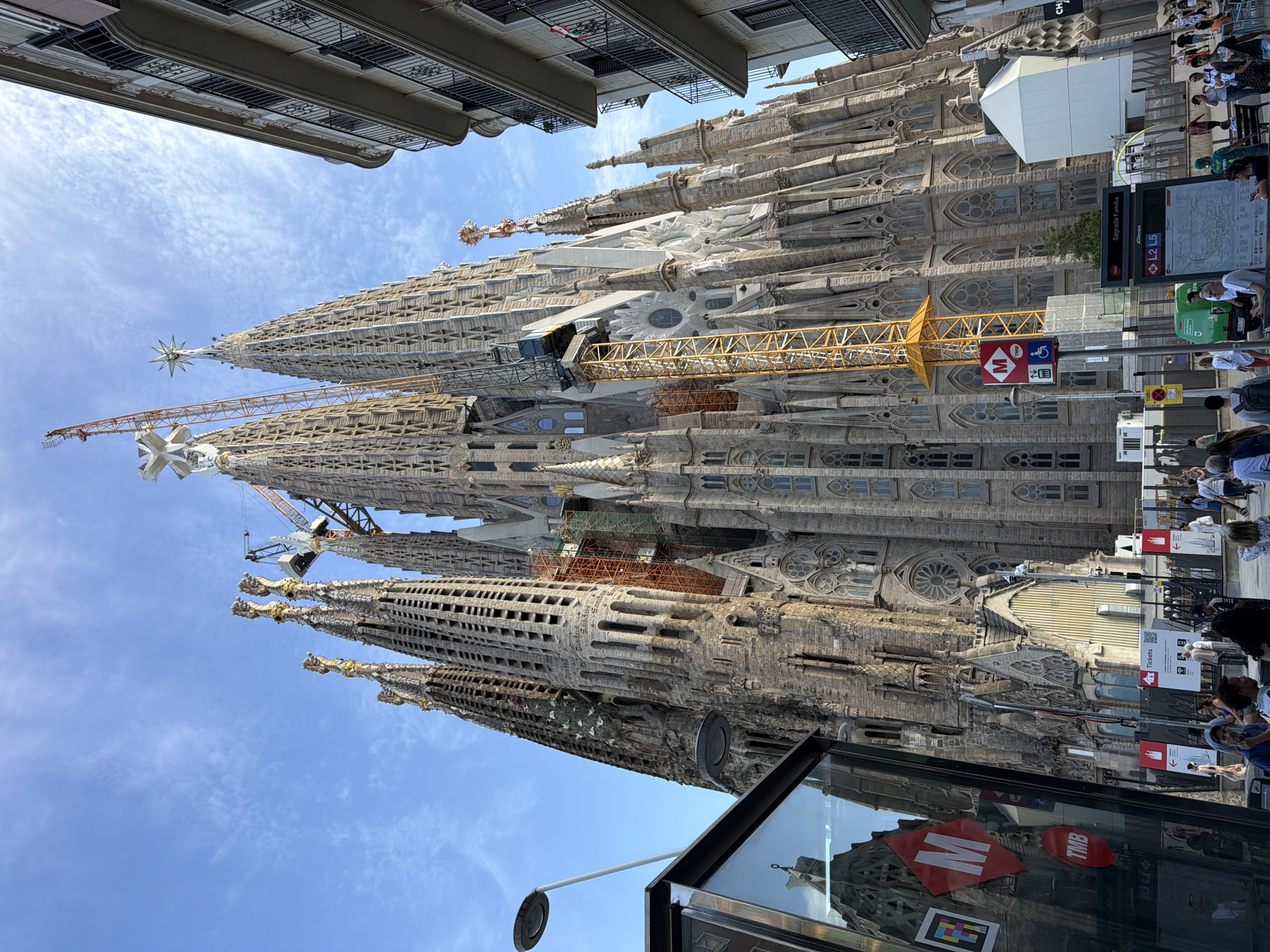 The image depicts the Sagrada Familia basilica in Barcelona, Spain. The basilica, a UNESCO World Heritage site, is a large, unfinished Roman Catholic church. The facade is characterized by intricate stone carvings and tall spires. Several construction cranes are visible, indicating ongoing work on the building. The sky is blue with scattered clouds. In the foreground, there is a glass enclosure with various stickers and signage, and a streetlamp. People are present in the scene, walking and standing, suggesting a public area. The image is captured from a low angle, emphasizing the scale and grandeur of the basilica.
