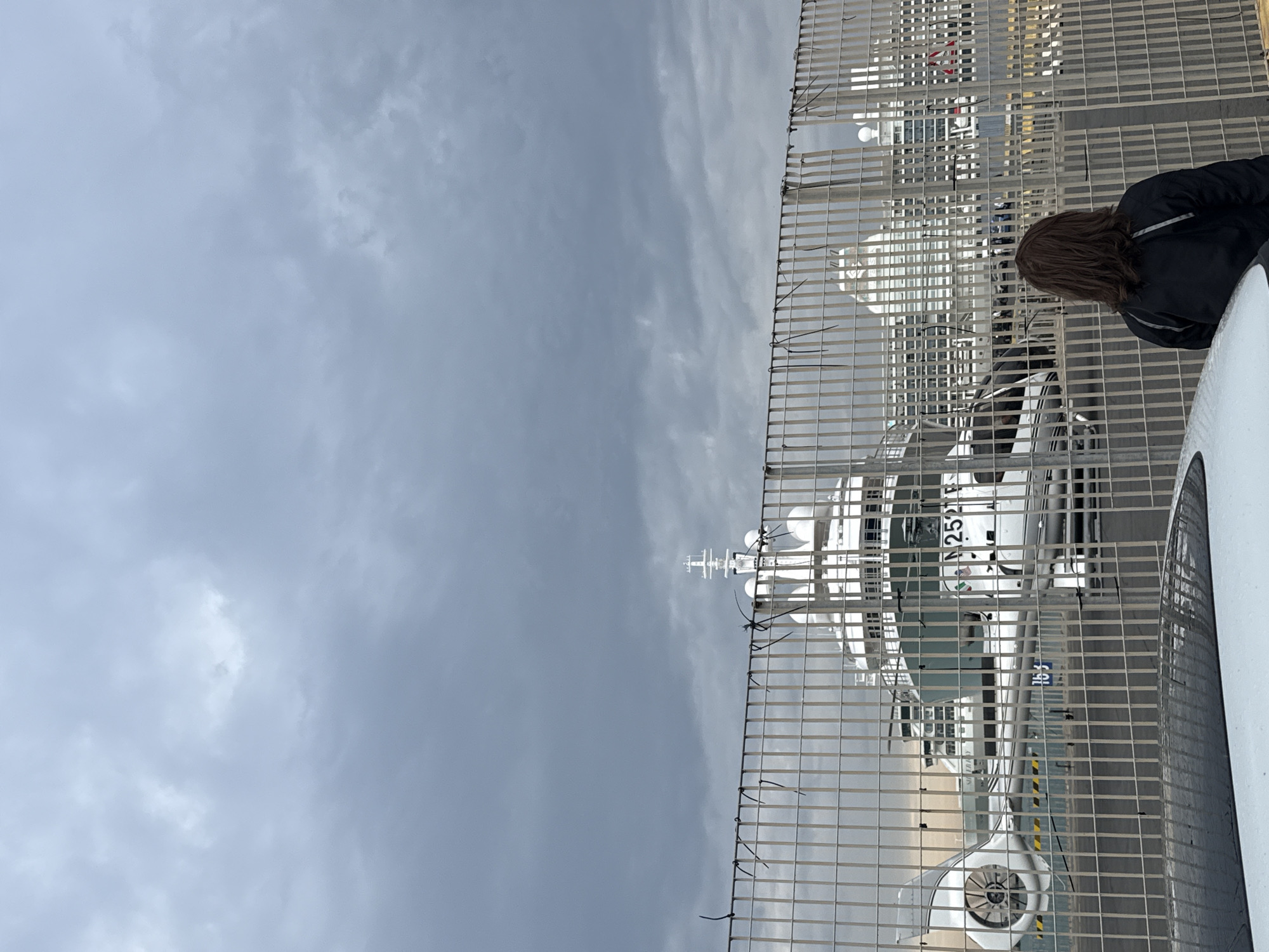 The image shows a harbor scene in Civitavecchia, Italy, as viewed through a chain-link fence. A white ferry or cruise ship, partially visible to the right, suggests a passenger port. Behind the fence is a helicopter with the marking "2521F". Part of a person in a black jacket is visible near the right edge of the frame. The sky is overcast with grey clouds.
