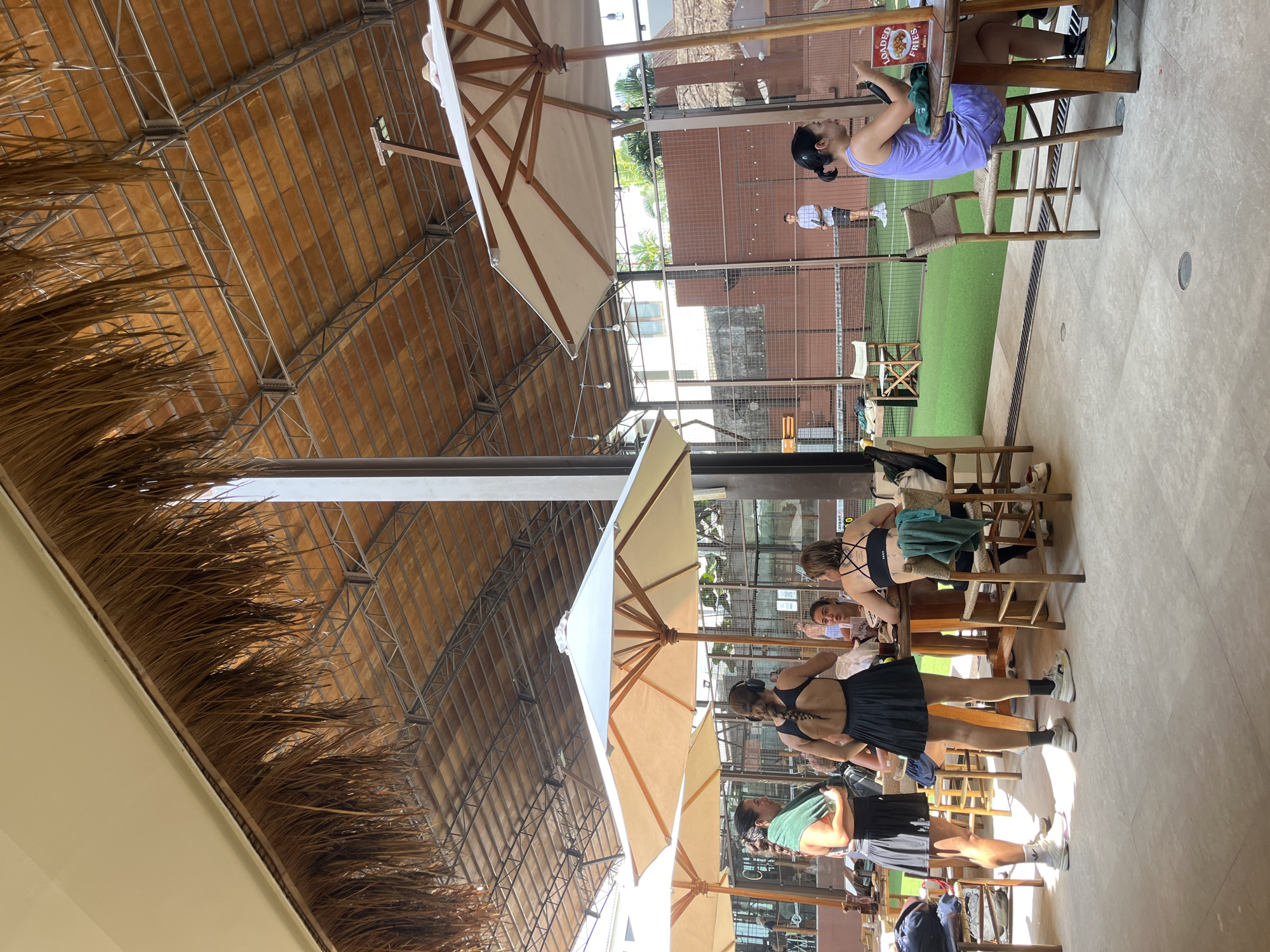 The scene is an open-air dining area in North Kuta, Indonesia. The location features a thatched roof supported by a steel frame, beneath which are several tables with seated customers. There are white umbrellas providing shade to the tables. Patrons, wearing casual attire, are seated at the tables engaging in conversation. Through a glass wall is a tennis court, where two people are standing.
