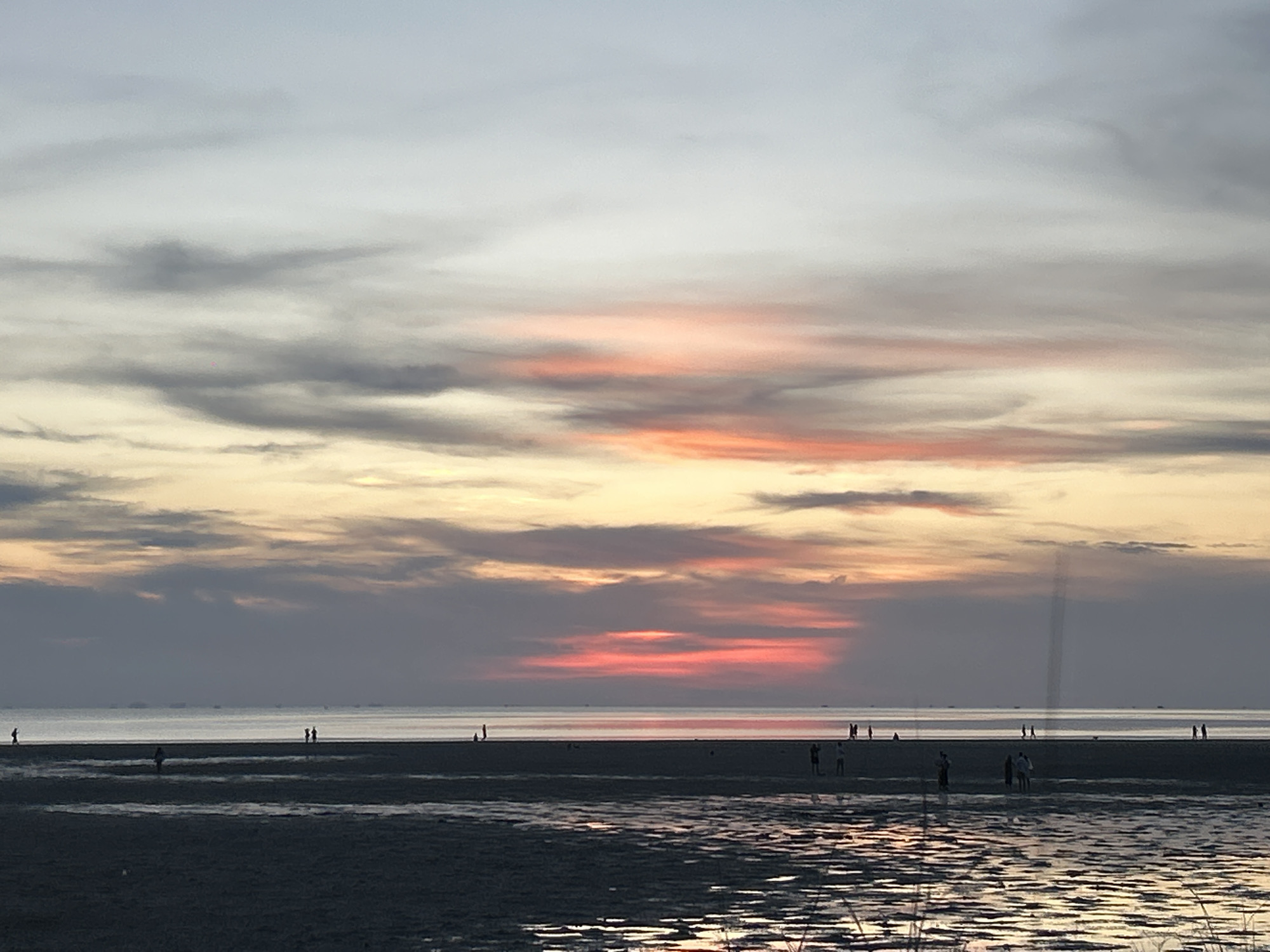 The image depicts a sunset over a beach in Baan Sri Thanu, Thailand. The sky is filled with clouds of varying shades of gray, orange, and pink, reflecting the setting sun. Several people are silhouetted against the water, walking along the shoreline. The tide appears to be low, leaving exposed wet sand. There are some unidentified objects visible in the distance near the horizon.