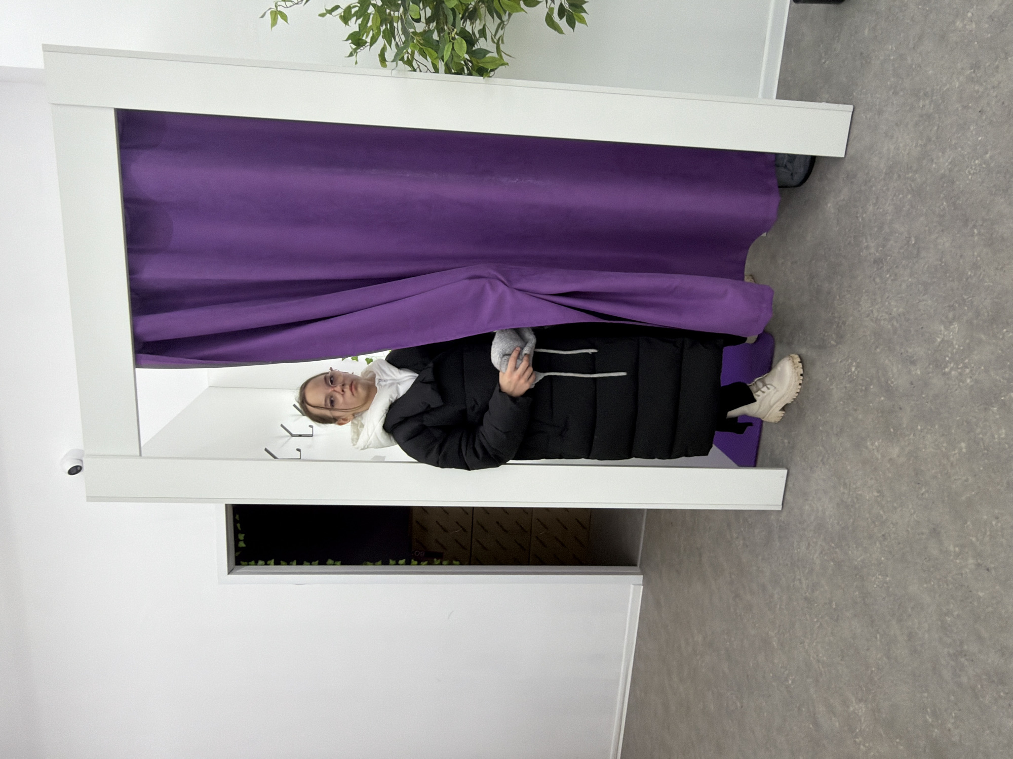 The scene is an indoor retail or studio environment located in Chaikovsky, Russia. A young female, positioned centrally, is partially visible as she emerges from a changing booth. She wears a long black puffer coat, a white hooded top, light-colored chunky-soled boots, and pink-framed glasses. In her hands, she holds a grey knitted item with white drawstrings. The changing booth consists of a white rectangular frame and a vibrant purple curtain, which is partially open. Inside the booth, three silver coat hooks are mounted on the white wall, and dark-colored footwear is visible on the floor beneath the curtain. To the left of the booth, an open doorway reveals a darker area with green artificial foliage and stacked brown boxes. A white security camera is mounted on the white wall above the left side of the booth frame. To the right, a potted artificial plant with green leaves is partially visible. The floor is a light grey, mottled surface.