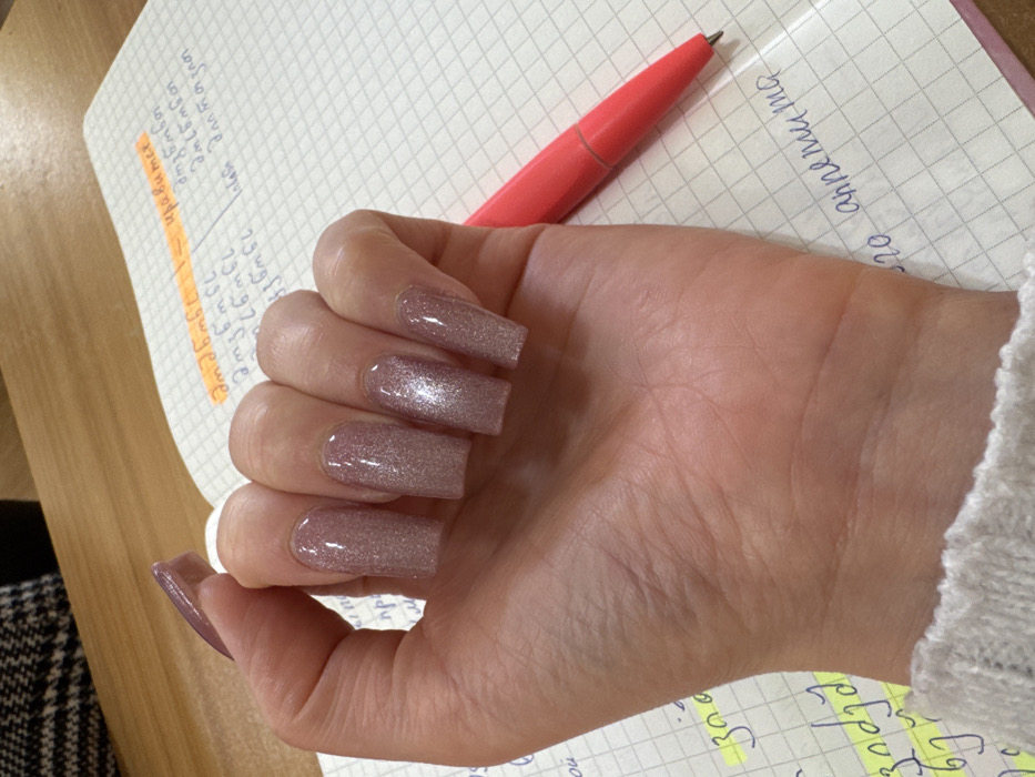 The scene depicts an indoor, close-up view of a person's left hand resting on an open notebook, likely a study or work environment in the city of Unknown, Unknown. The hand has light-toned skin, with fingers slightly curled and palm facing upwards. All visible fingernails are long, rectangular, and covered in a sparkling light pink or nude gel polish. A white, possibly knitted, cuff of a garment is visible around the wrist. The notebook features grid-lined paper with handwritten text in what appears to be Georgian script, some highlighted in yellow, and Cyrillic script words such as "нравится" and "аппетита". A bright red ballpoint pen with a visible tip lies horizontally on the notebook, to the right of the hand. The notebook and hand are positioned on a light brown wooden surface. In the upper left background, a portion of a dark, patterned fabric, possibly plaid or tweed, is visible.