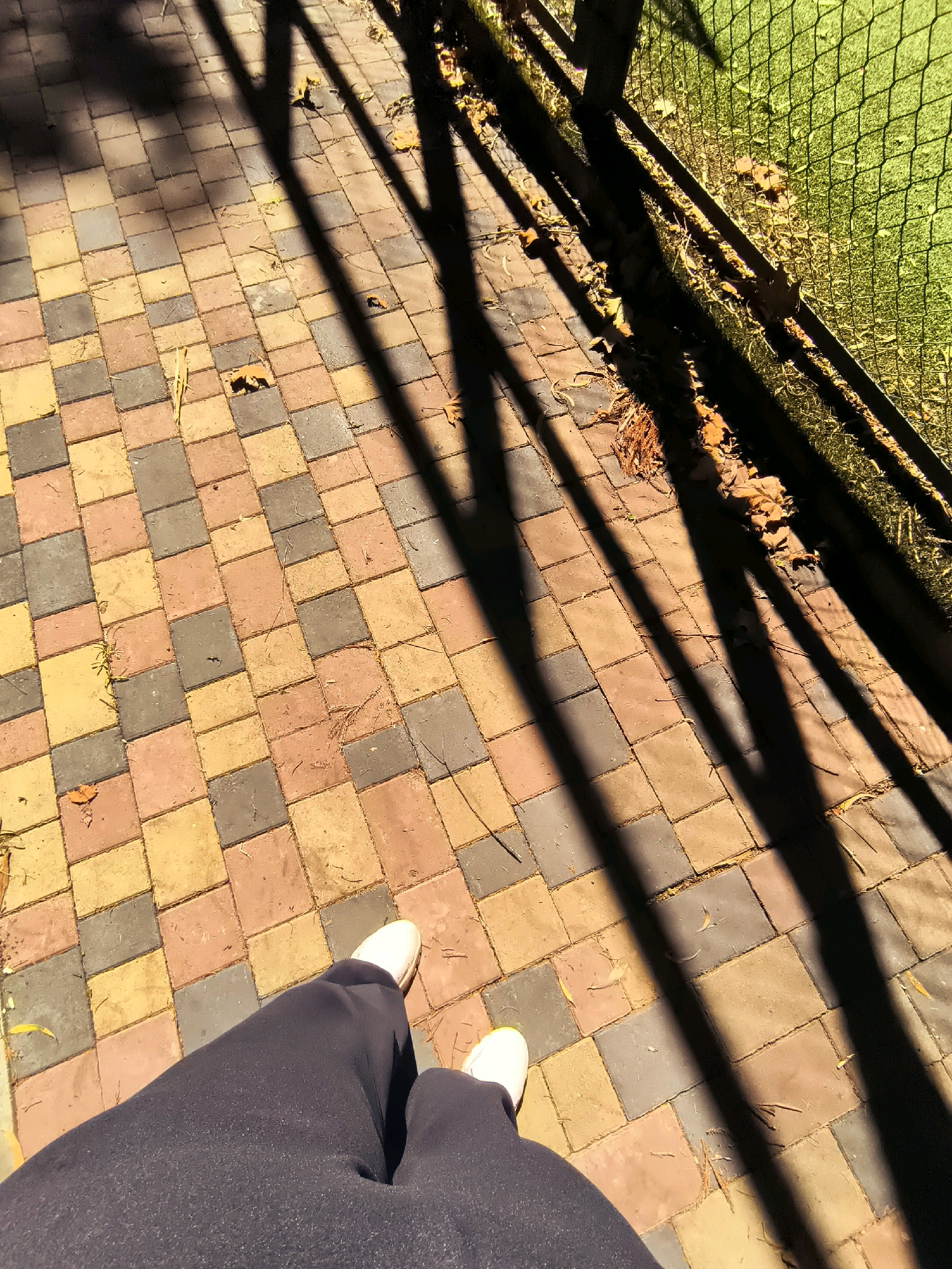 The image captures a ground-level perspective of a person's legs and feet, clad in dark athletic pants and white sneakers, standing on a paved walkway. The walkway is constructed with multi-colored square pavers in shades of orange, yellow, and gray, arranged in a repeating pattern. The ground is partially covered with fallen leaves and twigs, suggesting an outdoor environment, likely in autumn. Long, dark shadows from a fence or railing cast diagonal lines across the pavers. To the right of the walkway, a green grassy area is visible, bordered by a wire fence. The overall scene suggests a park, courtyard, or pathway in an urban or suburban setting. The image does not provide specific visual cues to pinpoint the location in Khelvachauri, Georgia, beyond it being an outdoor paved area.