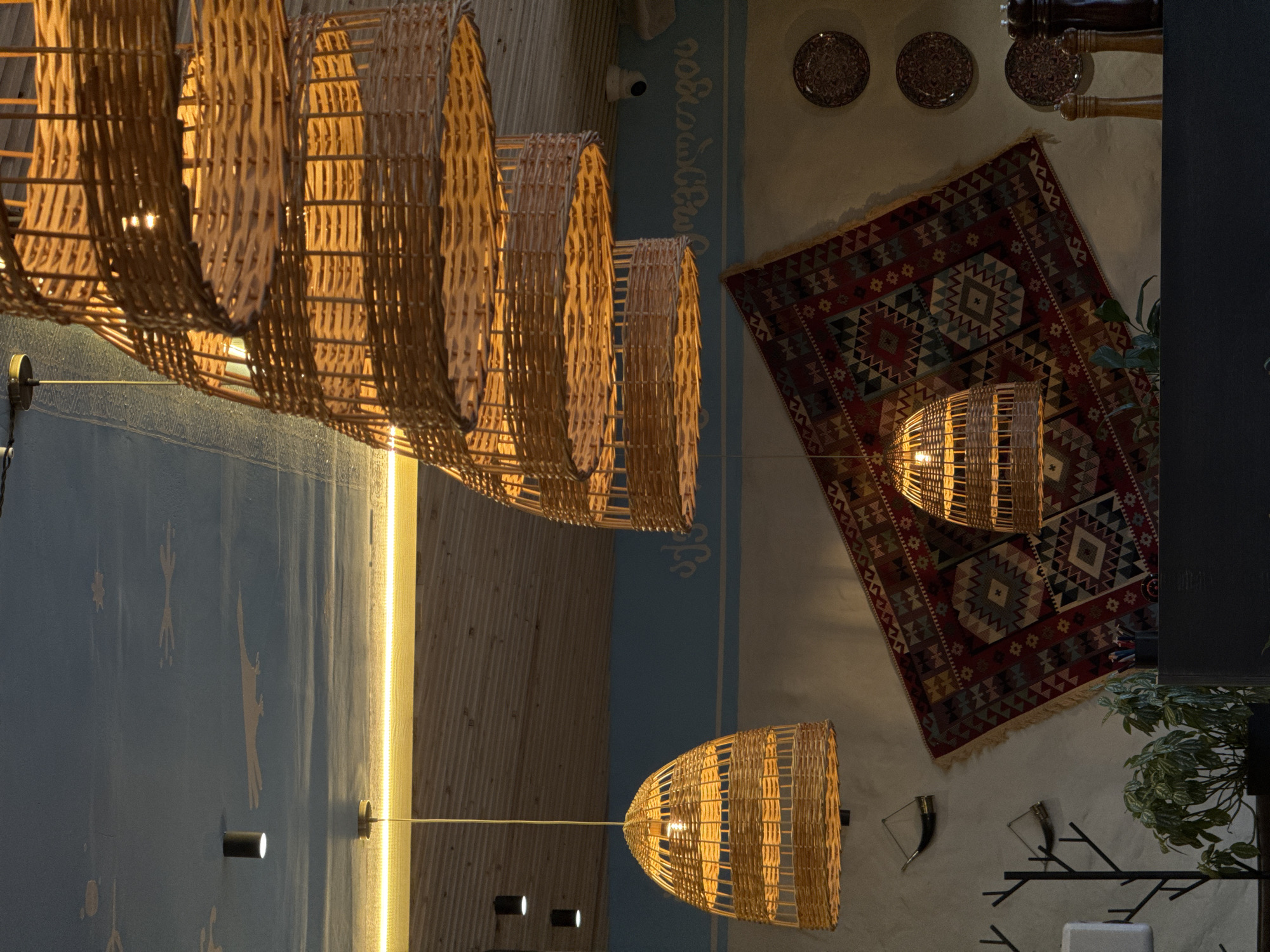 The image depicts an interior scene, possibly a cafe or restaurant, with a rustic and warm aesthetic. The focal point is a cluster of large, woven pendant lamps made of natural materials, casting a soft, amber glow. These lamps are suspended from the ceiling, which is adorned with a painted mural featuring abstract shapes and a faint image resembling a bird or perhaps a stylized cloud formation. A strip of warm LED lighting runs horizontally along the edge where the ceiling meets the wall.
Below the ceiling, wooden beams are visible, contributing to the natural and textured feel of the space. The wall behind the lamps features a striking, colorful tapestry or rug hung diagonally. This textile has a geometric pattern with various diamond and zigzag motifs in shades of red, blue, green, and cream.
To the left of the tapestry, two horn-shaped decorative objects are mounted on the wall. Further down and to the left, a dark, branch-like metal stand is visible, with a light-colored object attached to one of its branches. A potted plant with green foliage sits in the lower-left corner of the frame.
On the right side of the tapestry, three decorative plates with intricate patterns are mounted on the wall. In the foreground, a dark surface, likely a counter or table, runs across the bottom of the image. On this surface, a set of salt and pepper grinders and a small pot of utensils are visible.
The overall impression is one of a cozy and artistically decorated space. While the image itself does not contain any people or explicitly indicate a specific city, if this scene were located in Mikhaylovsk, Russia, it would suggest a place that values decorative arts and a natural, inviting ambiance.