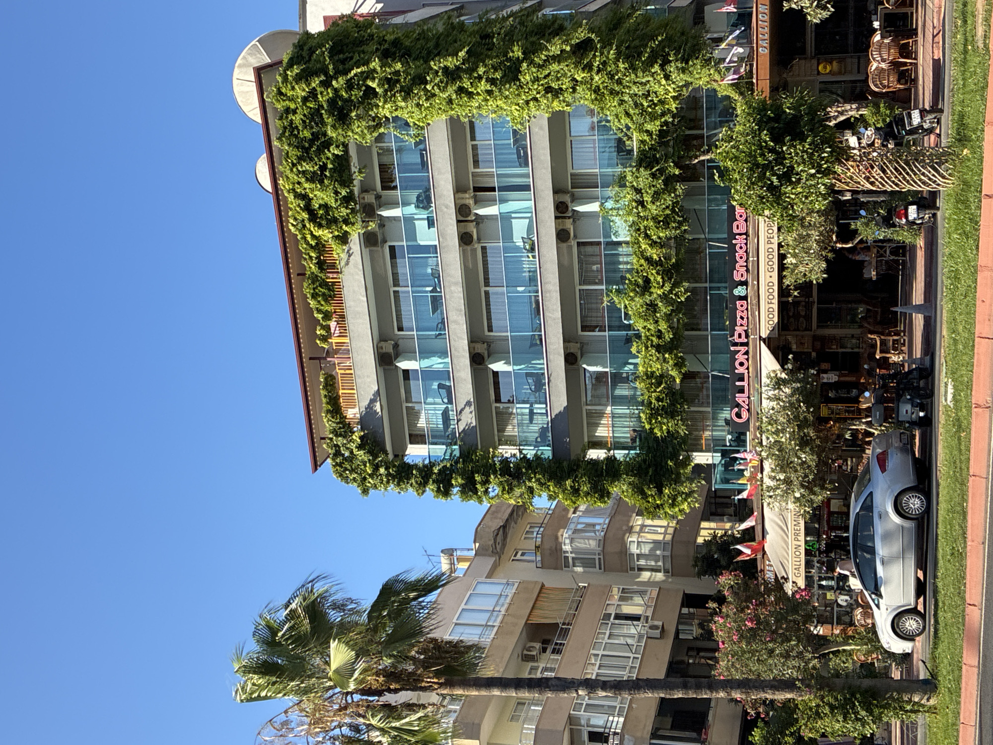 The image shows a street scene in Alanya, Turkey, featuring a multi-story building with lush green vines growing over its facade. The building has multiple balconies with glass railings and outdoor seating areas. In front of the building, there is a street with a silver car parked, a palm tree, and vegetation. A neon sign above a storefront reads "GALLION Pizza & Snack Bar" and below it, another sign says "GOOD FOOD • GOOD PEOPLE". Several flags are displayed on poles in front of the establishment. The sky is clear and blue. Another building, taller and lighter in color, is visible to the left of the main building. The ground in front of the buildings appears to be a sidewalk with outdoor seating and tables for patrons. Two motorcycles are parked on the sidewalk.