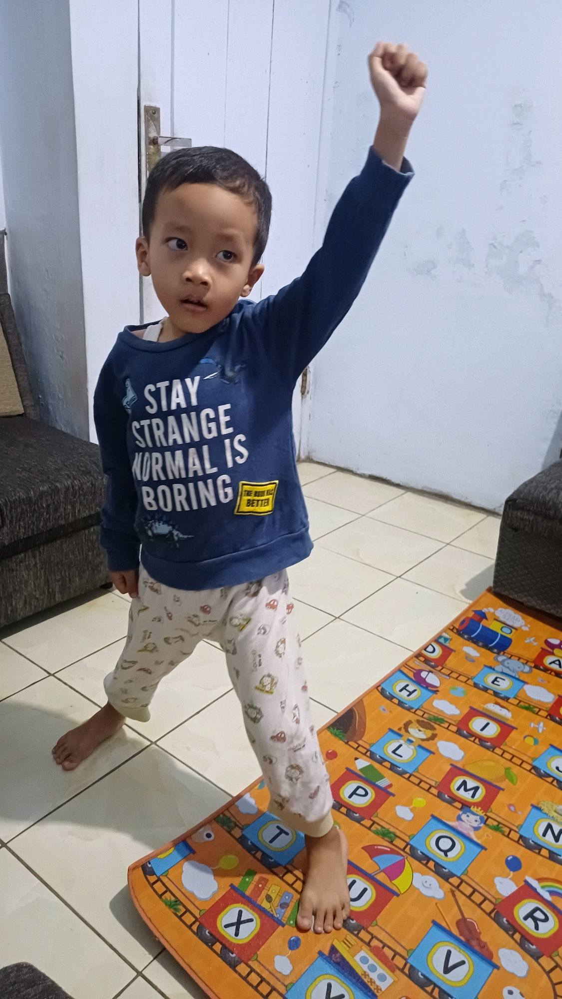 A young boy stands on a mat with letters and trains printed on it. He is wearing a dark blue sweatshirt with the words "STAY STRANGE NORMAL IS BORING" printed in white letters, and a yellow patch that says "THE BODY KILL BETTER". He is also wearing light-colored pajama pants with a pattern of animals and objects. The boy's right arm is raised in the air with his fist clenched. He is looking to his left, with his eyes slightly unfocused. The mat is orange and features a colorful alphabet train design. The floor appears to be tiled. The background shows a white door and a wall with some discoloration. This scene is located indoors, within a residential setting in Kowangan, Indonesia.