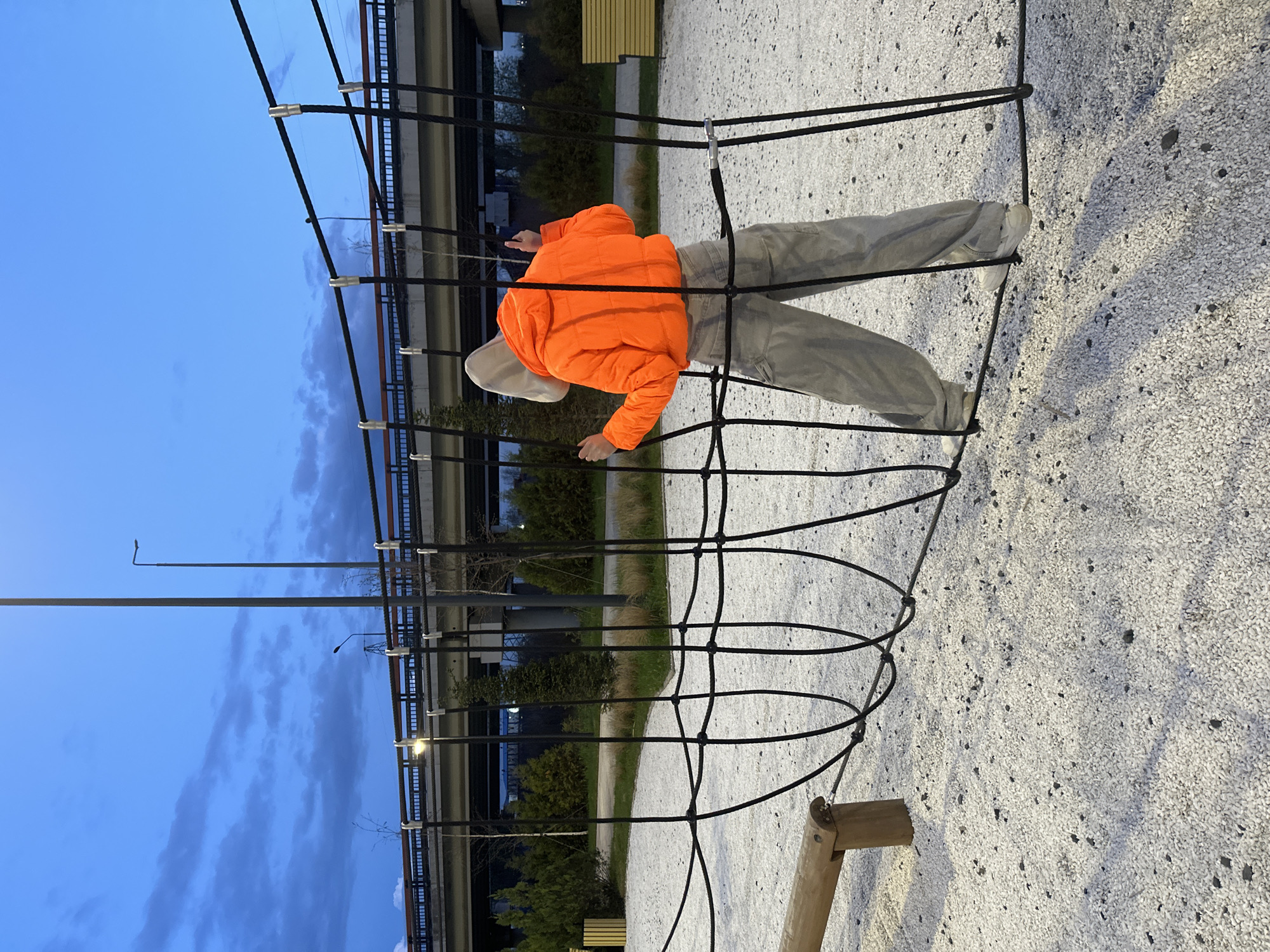 A person wearing an orange jacket, light-colored pants, and sneakers is climbing a black rope structure in a playground in Rostov-on-Don, Russia. The playground has a light-colored gravel surface. In the background, there is a highway overpass. The sky is a gradient of blue, suggesting it is dusk or dawn.