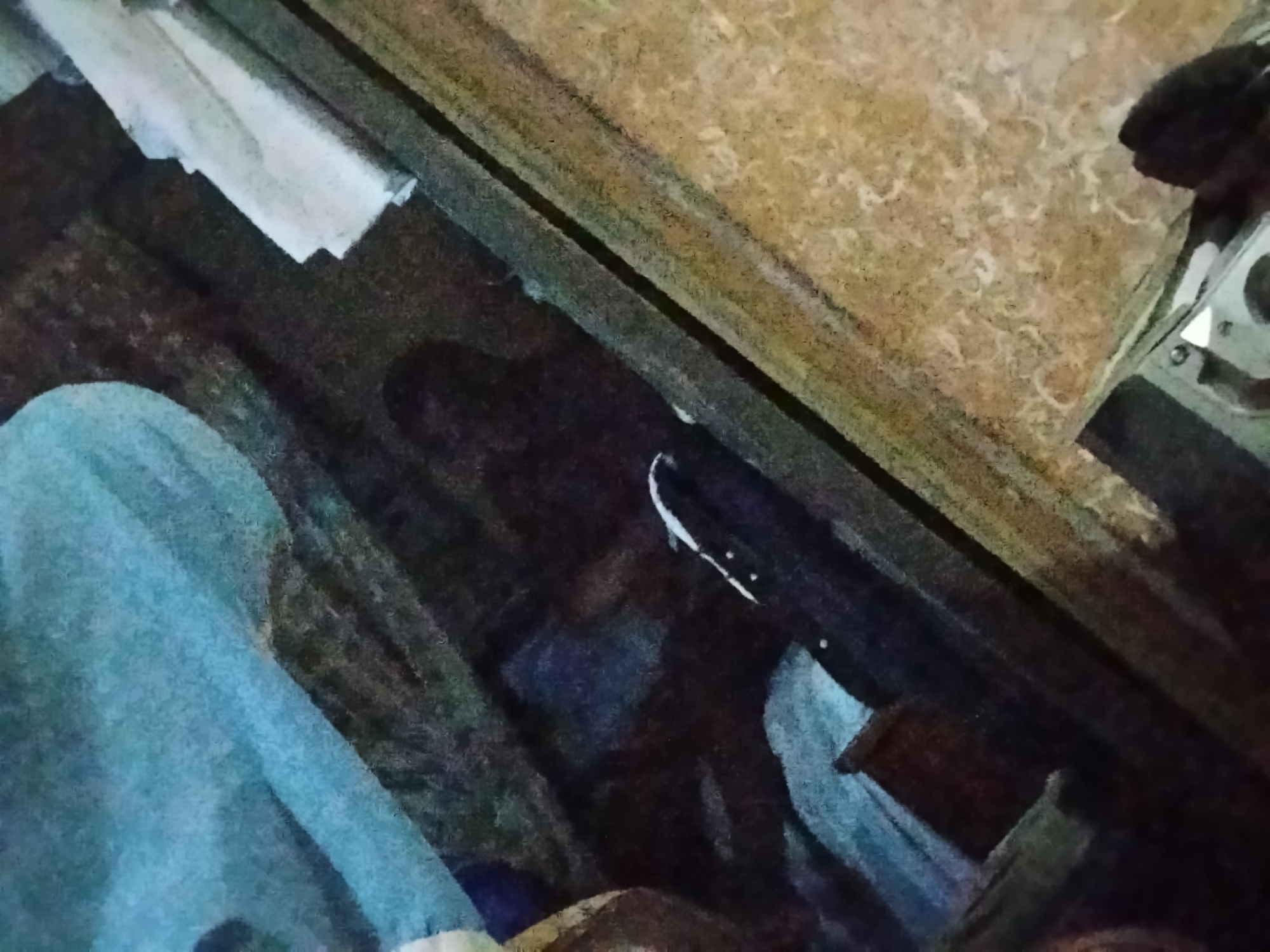 The image shows a cluttered interior. A person is visible in the lower-left, covered with a light blue cloth. A wooden structure or piece of furniture with a marbled top is present on the right. A dark object with a white line and rivets sits in the center. It's difficult to determine specifics about location or activities based solely on this obscured image. There is no discernible information confirming its location in Shangisha, Nigeria.