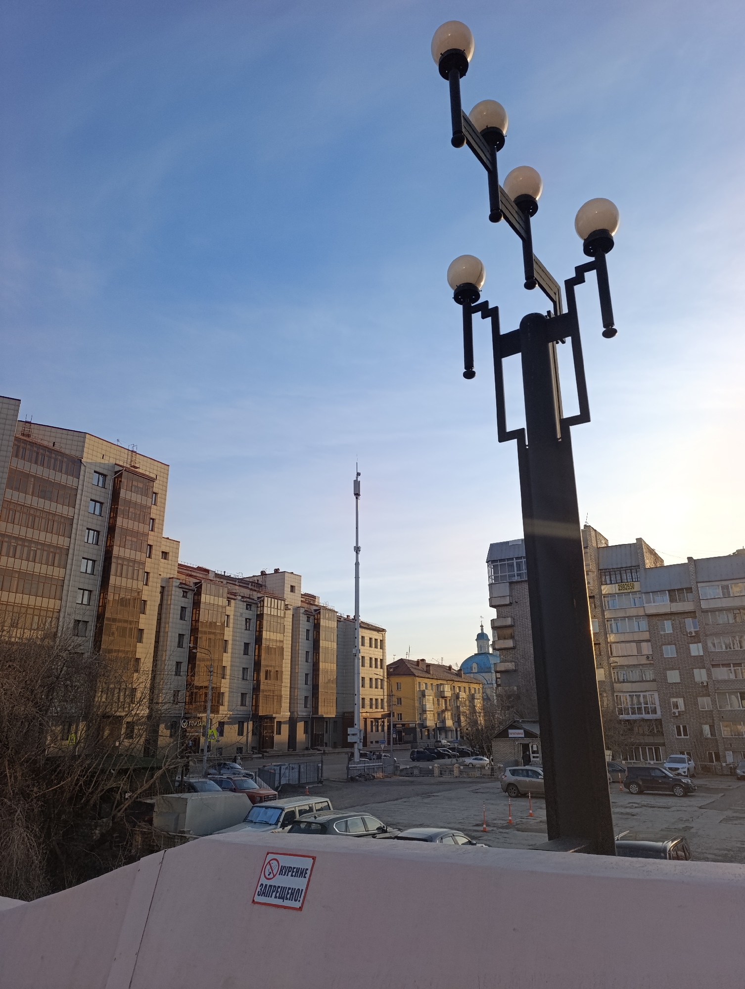 This image depicts an urban street scene in Krasnoyarsk, Russia, likely captured during the early morning or late afternoon due to the low sun angle casting warm light on buildings and creating lens flare. The sky is a clear, bright blue with minimal cloud cover.
In the foreground, a distinctive dark streetlamp, featuring a stylized geometric design with five spherical white light fixtures, stands prominently on the right. A light-colored concrete barrier forms the bottom edge of the frame, on which a white sign with red lettering and a prohibition symbol is affixed. The sign reads in Russian: "КУРЕНИЕ ЗАПРЕЩЕНО!" (SMOKING FORBIDDEN!).
Beyond the barrier, a parking area filled with various cars is visible. On the left, bare trees and bushes suggest a colder season. The background is dominated by several multi-story buildings, appearing to be a mix of residential and commercial structures. Some buildings feature modern designs with large, reflective glass windows, while others have more traditional stone or brick facades.
A tall, slender communications or cellular tower rises high into the sky in the mid-ground, serving as a noticeable vertical element in the cityscape. Further in the distance, a light blue church dome with a spire and cross can be seen, adding a historical architectural detail. One building in the background also displays the text "2592650" (likely a phone number), and a small white building has "МАГАЗИН" (STORE) written on it. No people are visible in the scene, which conveys a tranquil, everyday urban atmosphere.