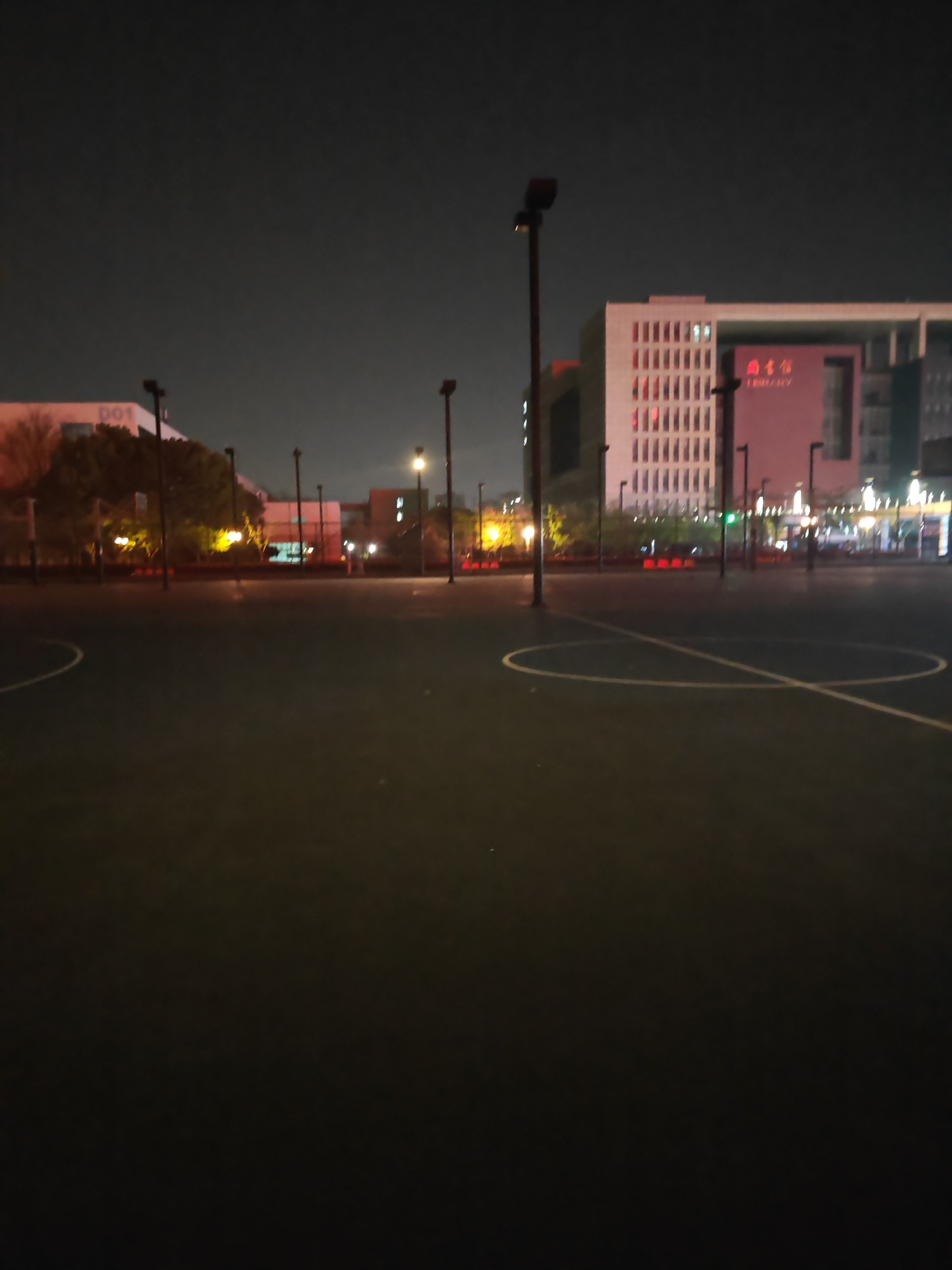 The image depicts an outdoor basketball court located in Jiangning District, China. The court is surrounded by tall light poles and the court's surface appears to be a dark color. In the background, there are several buildings, one with the word "LIBRARY" illuminated. The sky is dark, suggesting it is night. The scene is devoid of people.