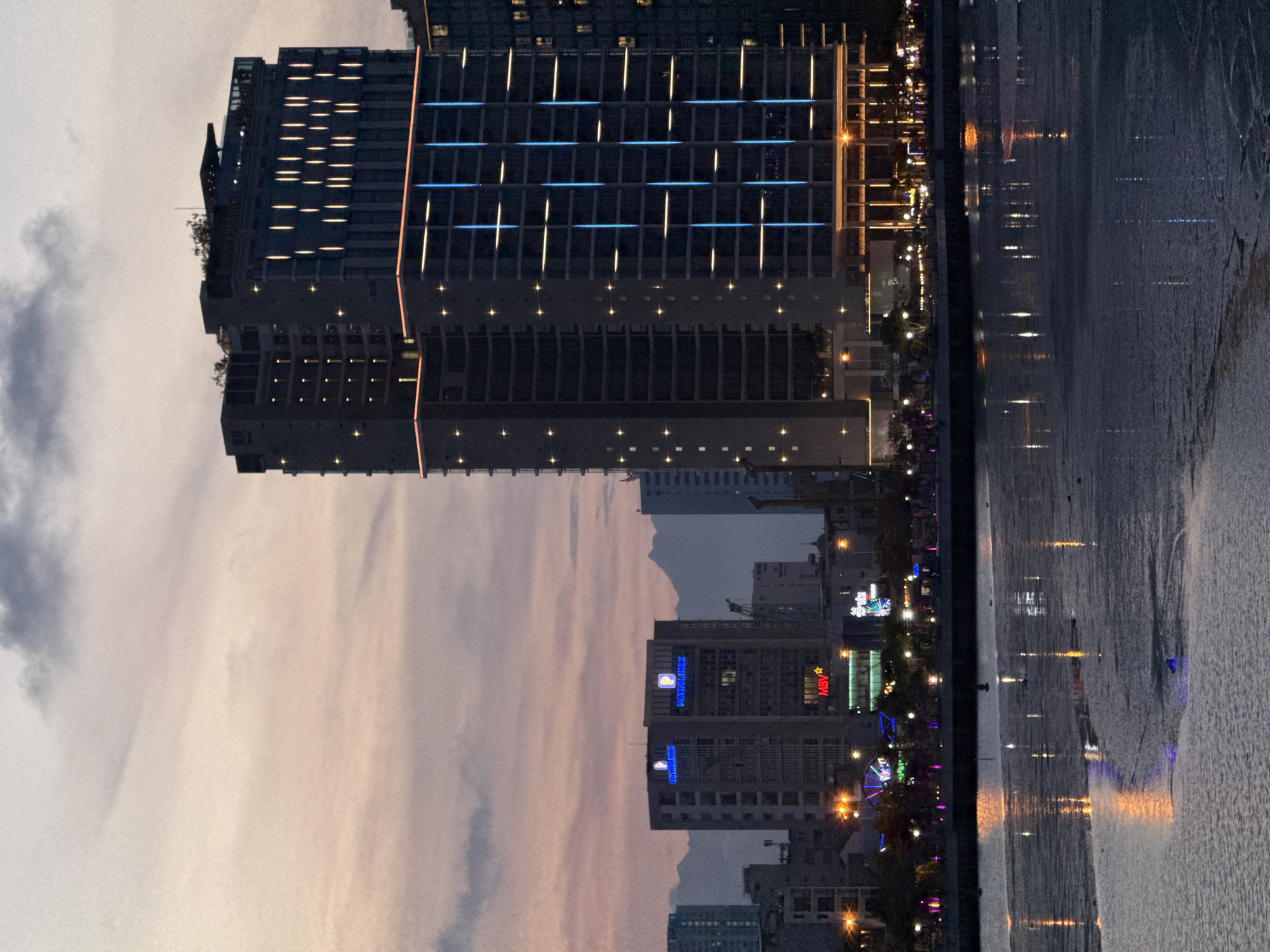 This is a vertical, outdoor, low-angle shot taken at dusk in Da Nang, Vietnam. The scene features a body of water with reflections of buildings and lights, and a cityscape on the far side. The water occupies the right side of the frame, showing ripples and colorful reflections from lights.
Dominating the left and center of the image is a large, modern building with a facade of windows. Some of these windows are illuminated with bright yellow and blue lights, creating a grid-like pattern. The building appears to be a hotel or office tower, with tiered sections and some greenery visible on balconies or setbacks.
Further into the cityscape, other buildings are visible, some with illuminated signs and windows. One prominent sign displays the text "MBV" in red letters. Another building has a blue illuminated sign that reads "POSTEDAMINO" with a yellow logo above it. A Ferris wheel with colorful lights is also visible in the distance, suggesting a recreational area or amusement park.
The sky is overcast with streaks of pink and orange, indicative of sunset. The overall mood is calm and illuminated by the city lights and reflections on the water, creating a serene urban night scene. No people are directly visible in the frame.