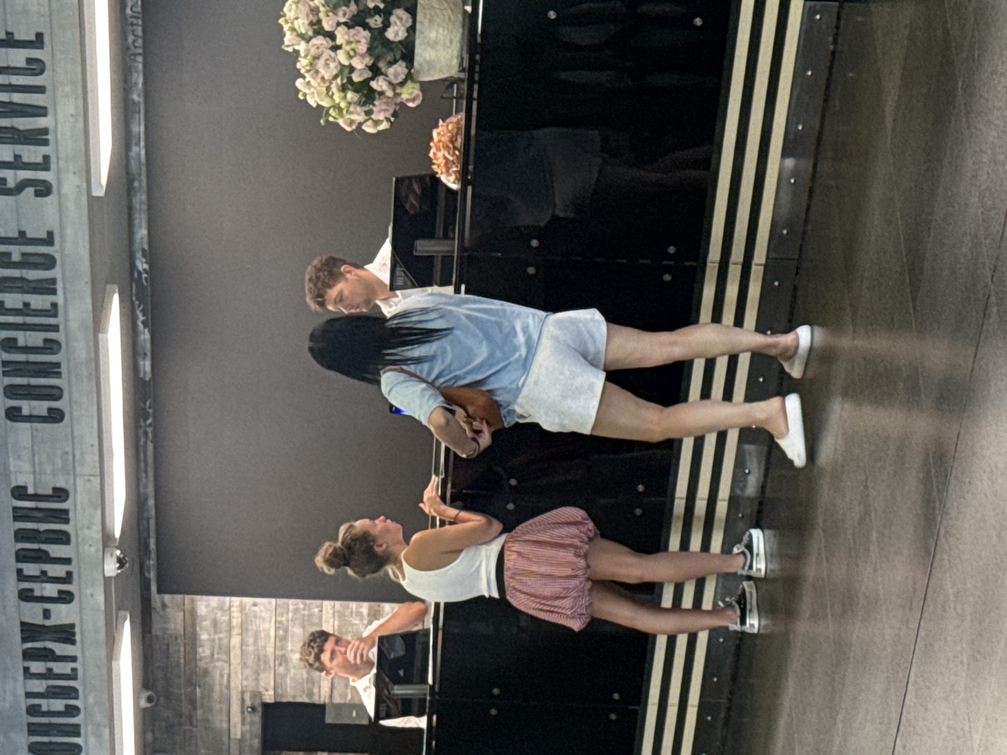 A man and a woman are standing at a concierge desk in what appears to be a hotel lobby in Yalta, Ukraine. The man, wearing a white polo shirt, is seated and looking at a laptop. The woman, wearing a white tank top and a striped skirt, is standing and facing away from the camera, with her hands on the desk. Another woman, wearing a blue top and white shorts, is also standing at the desk, facing the man. A floral arrangement is visible on the desk to the right, along with some small food items. The concierge desk is dark and glossy, with decorative stripes along its base. The background features a textured wall and a sign that reads "КОНСЬЕРЖ-СЕРВИС CONCIERGE SERVICE". Security cameras are mounted on the ceiling. The floor is a reflective gray tile.