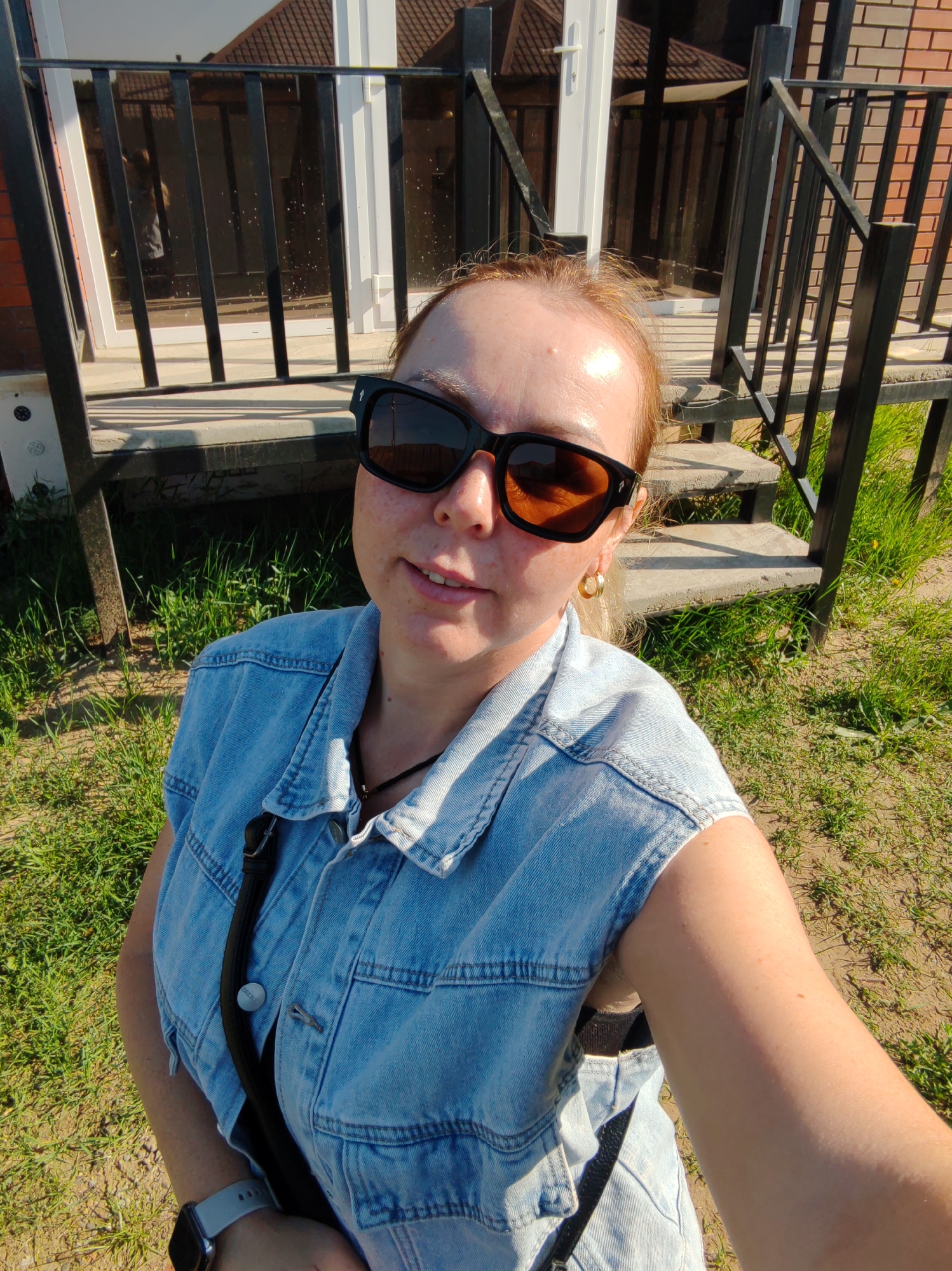 A woman stands in front of a house with a small porch in the city of Chaikovsky, Russia. The woman has red hair, fair skin, and is wearing sunglasses. She is wearing a blue denim vest and a black necklace. A black strap crosses her body. She is wearing a smartwatch on her left wrist. The house has a brown brick facade and black metal railings on the porch. Green grass and bare earth are visible in the foreground.