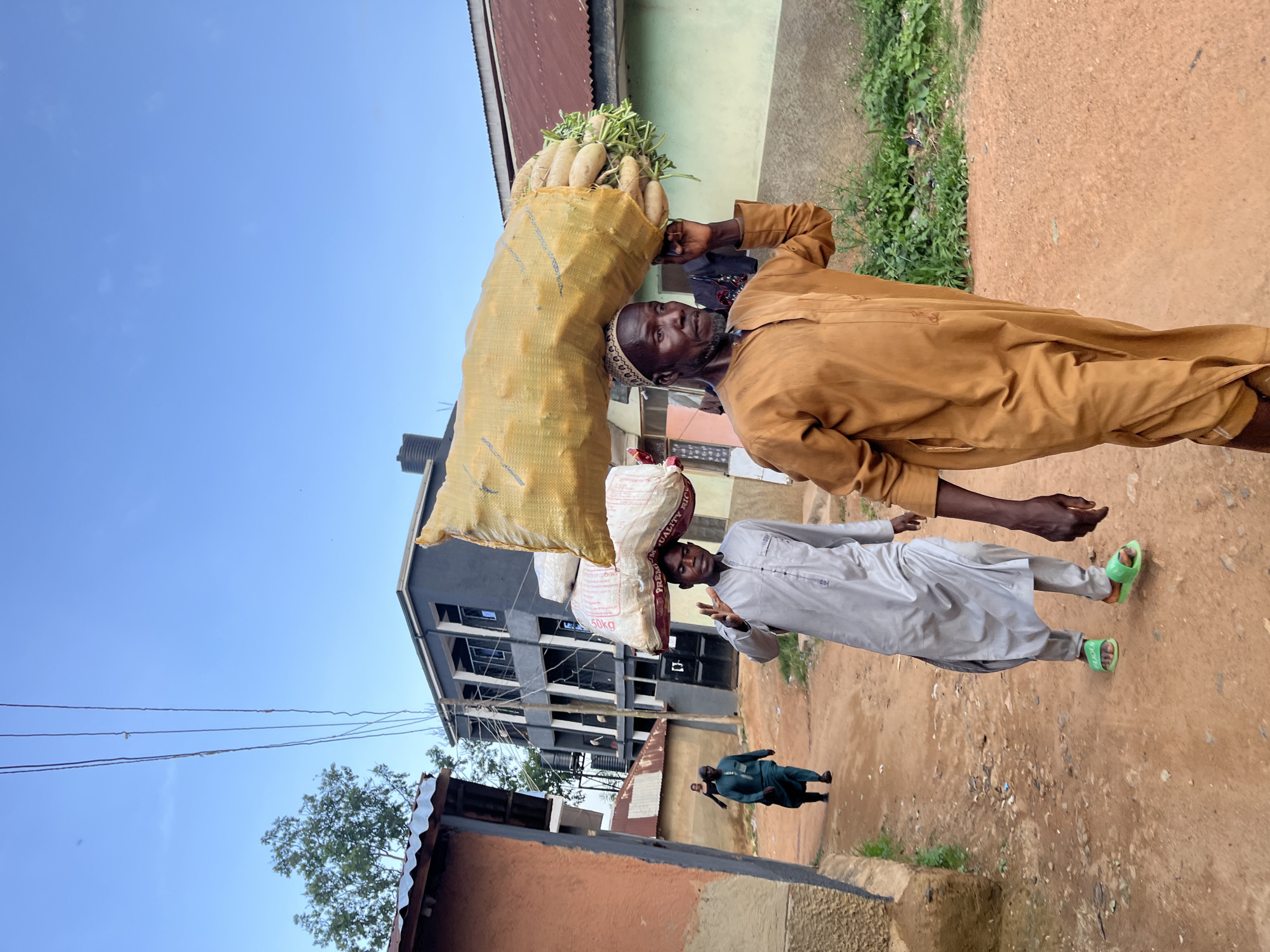 In Rafin Par, Nigeria, a man in a tan kaftan walks carrying a large, yellow sack filled with white root vegetables on his head. He is wearing a traditional cap. To his left, another man in a light gray kaftan also carries a load on his head, which appears to be two white sacks. He wears bright green sandals. Further behind them, a third man in a teal kaftan walks down the dirt path. The background shows a multi-story gray building with balconies and windows, typical of urban architecture in Nigeria, alongside smaller, rustic buildings with corrugated metal roofs. The path is unpaved and reddish-brown. Overhead, electrical wires crisscross the sky under a clear blue sky.