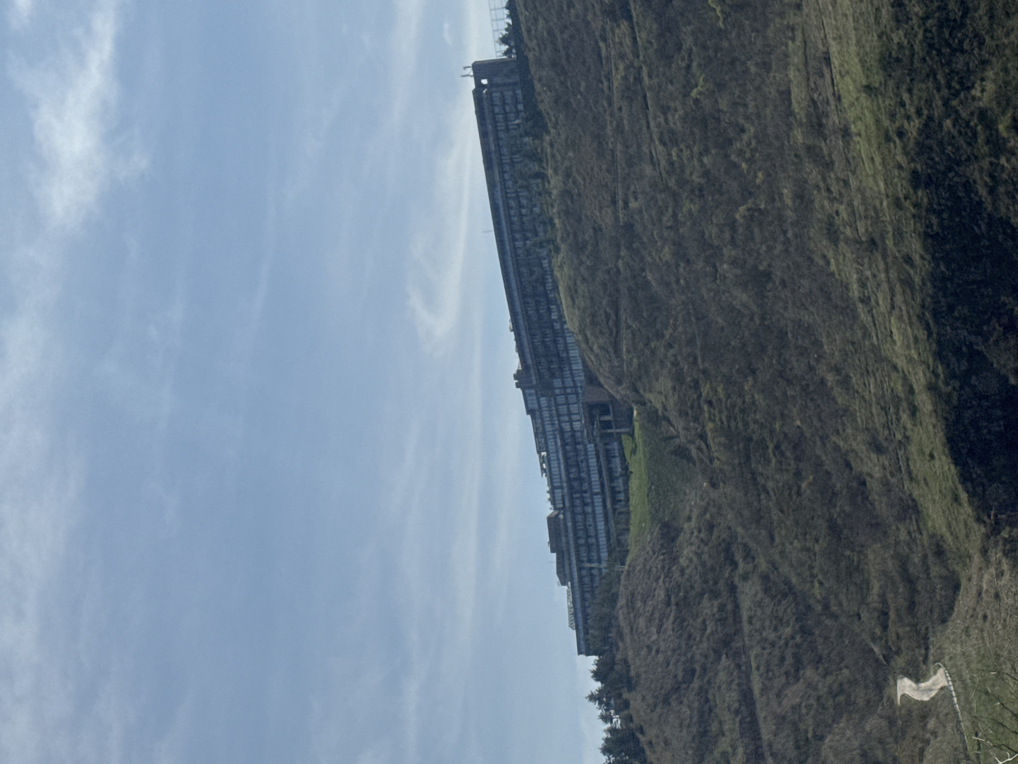 The image shows a long, multi-story building nestled against a hillside. The building's facade features numerous windows, suggesting it may be an office or apartment complex. The hillside is covered in vegetation, including trees and grasses, with varying shades of green and brown. The sky is visible above the building and hillside, with scattered clouds.
The building is the Tbilisi City Assembly building located in Tbilisi, Georgia.