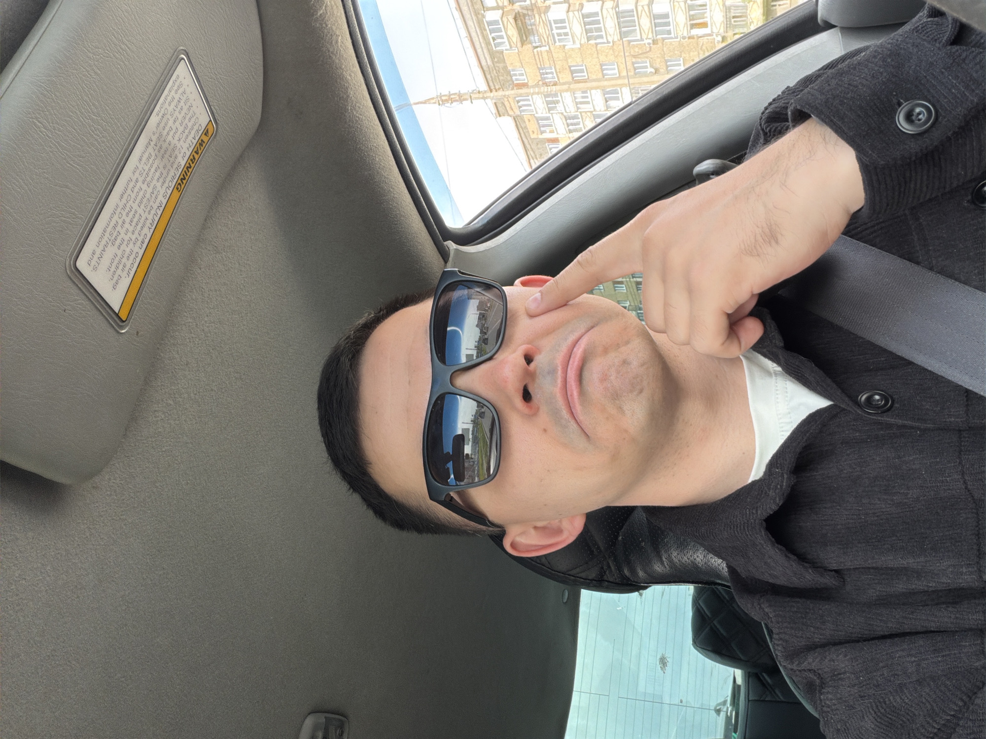 A selfie, taken inside a vehicle, depicts a person with short, dark hair wearing sunglasses, a white shirt, and a dark jacket. The person points to the skin between the sunglass frame and the eye. A seatbelt is visible. The car's interior includes a sun visor with a warning label. Through the window, a building with multiple windows is visible, reflecting in the sunglasses. The reflection suggests the location is in Izhevsk, Russia, based on distinct architectural features and building coloration.
