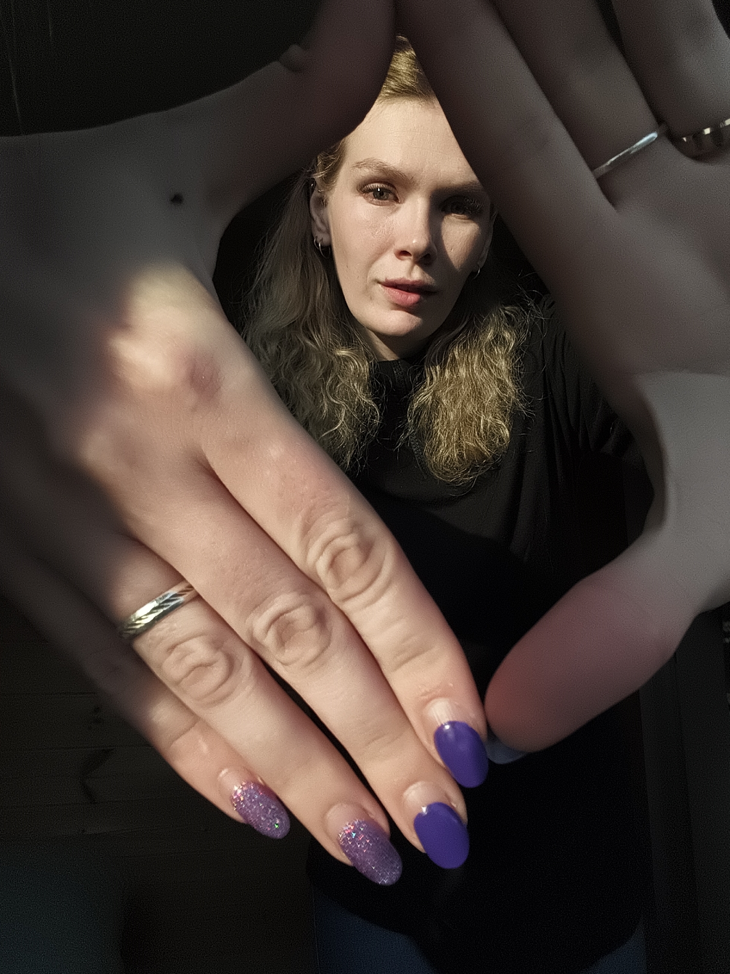 A person with light complexion, curly blonde hair, and wearing a black turtleneck shirt is centered and framed by two hands in the foreground. The left hand has a mole on the palm, a silver ring on the ring finger, and the fingernails are painted with a purple glitter polish. The right hand has two rings on the fingers and the fingernails are painted solid purple. The lighting is inconsistent, with the left hand in direct sunlight and the rest of the scene in shadows. The location, Русский Вожой, Russia, cannot be determined from this image.