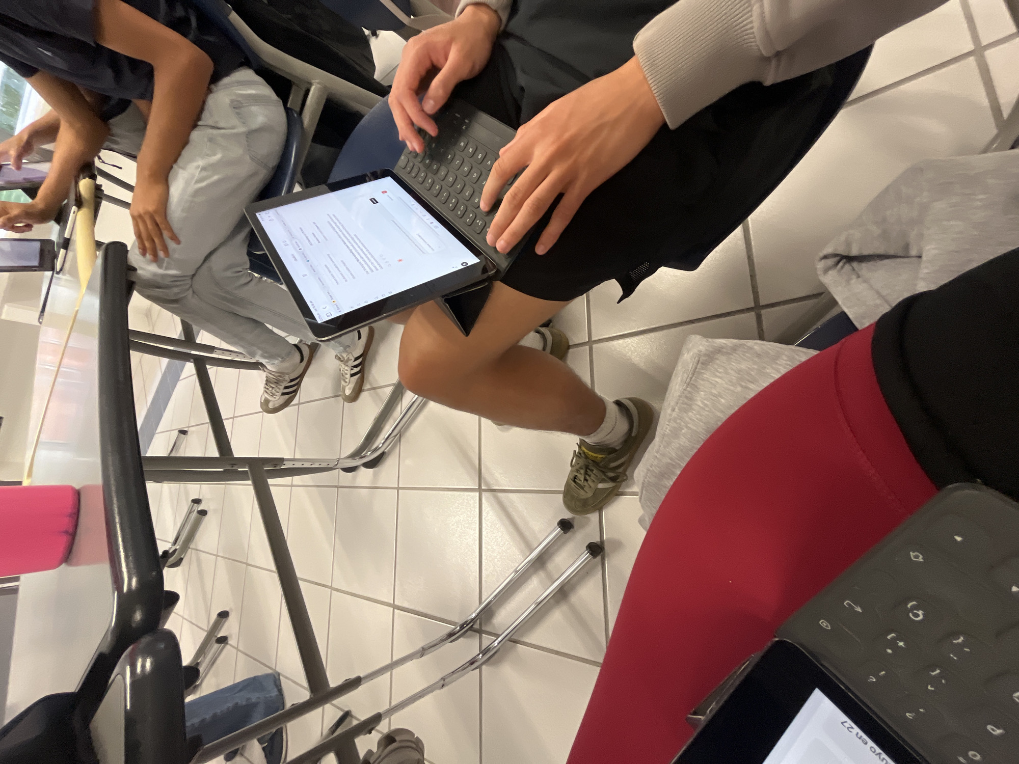 This media file captures an indoor scene, likely in a classroom or study area in Puebla, Mexico, showing several individuals engaged with electronic devices. The image is taken from a high angle, looking down at people seated around tables or desks.
The primary subjects are at least three people. In the center, an individual wearing black athletic shorts and a light grey long-sleeved top is actively typing on a tablet connected to a detachable keyboard, held on their lap. Their feet, clad in olive green sneakers and white socks, are visible on the tiled floor. To the left, another person wears light-colored jeans and a dark top, also appearing to interact with a device, though it's mostly obscured. This person wears white sneakers with black stripes. On the far right, partially visible, another person wears red leggings and a dark top, with a grey sweatshirt resting nearby. They also have a tablet with a keyboard, oriented differently.
The activity appears to be studying, working, or typing, as multiple individuals are focused on their tablets. The setting features white tiled flooring with grey grout lines, and metal-framed chairs and tables are scattered throughout the space. The lighting suggests an indoor environment, but the time of day is not discernible.
Notable details include the various sneakers worn by the individuals (Adidas-style on the left, olive green on the center person), and the technology being used, which consists of tablets equipped with external keyboards, suggesting more intensive typing or data entry than typical tablet use.
Visible text on the screen of the tablet in the center, when rotated, shows "uyo en 27". The same text fragment is also visible on the tablet to the right. The screen displays what appears to be a document or a text editing interface, with a search or input bar visible at the top. No other specific location cues beyond the general indoor setting are visible in the image itself.