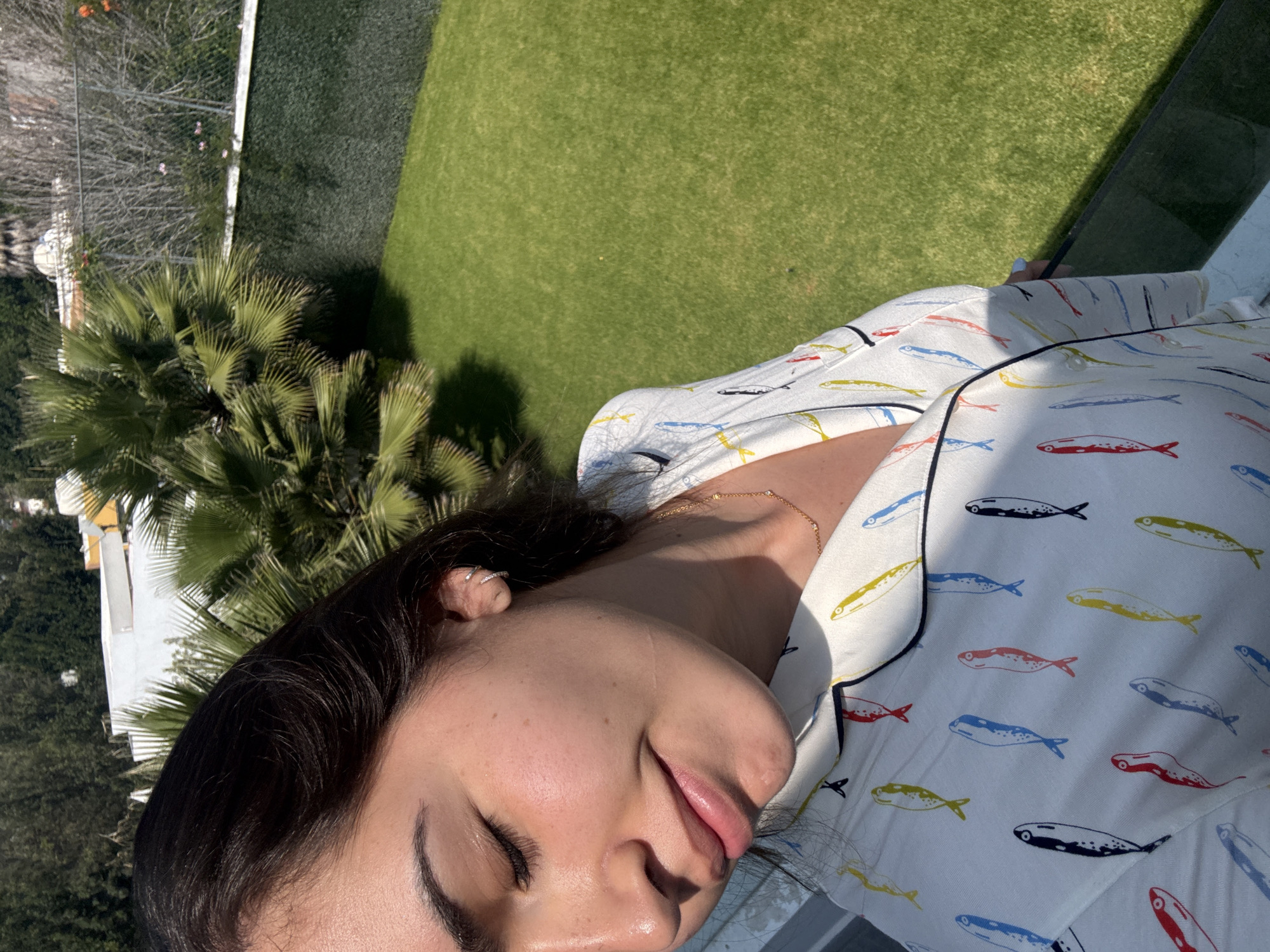 A close-up, rotated view captures a person, likely female, relaxing outdoors in bright sunlight. Her eyes are closed, and she wears a serene, slightly smiling expression, with dark hair framing her face. She has small silver hoop earrings and a delicate gold chain necklace. Her attire is a light-colored top, possibly pajamas, adorned with a repeating pattern of stylized fish in various colors, including red, blue, yellow, and black, accented with black piping on the collar.
The setting appears to be a balcony or terrace, offering a downward view of a lush outdoor landscape. The background features an expansive, well-maintained green lawn, suggesting a garden or golf course. Numerous vibrant green palm trees and other dense foliage are prominent, casting distinct shadows on the sunny ground. In the distance, bare trees and a white structure or fence are partially visible. A clear glass barrier is present in the foreground, separating the person from the distant view.
The image conveys a sense of peaceful relaxation under the bright daytime sun, as indicated by the strong highlights on the person's skin and the sharp shadows. No discernible text is visible within the image. The location context for this scene is San Bernardino Tlaxcalancingo, Mexico.