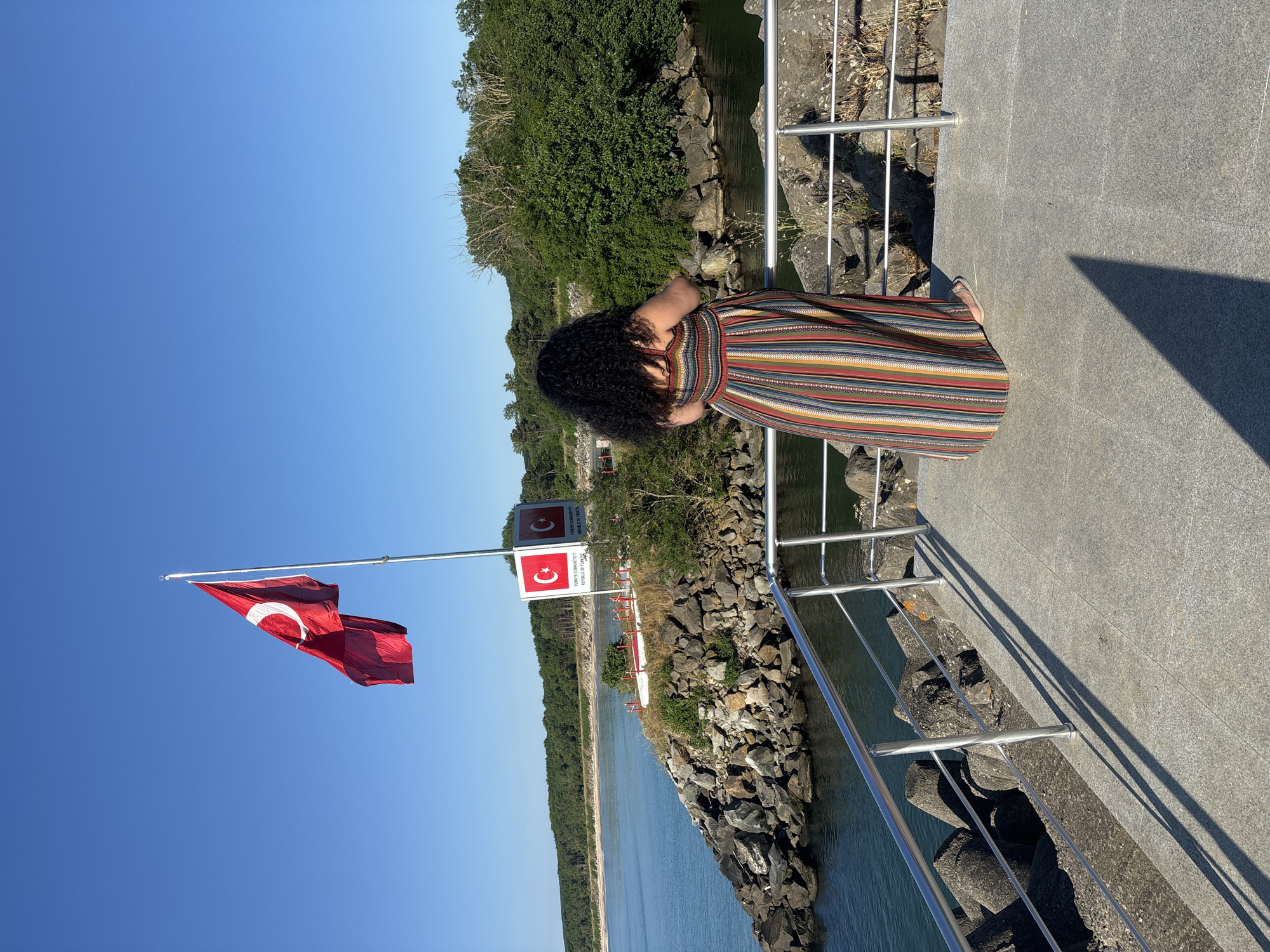 A woman with curly dark hair stands on a paved walkway, facing away from the camera, looking out at a body of water. She is wearing a long, striped maxi dress in various colors. The walkway is bordered by a metal railing. Beyond the railing, there is a rocky shoreline with large concrete wave-breakers. A Turkish flag flies on a flagpole on a rocky outcrop in the distance, next to a sign with Turkish flags and text that reads "TÜRKİYE CUMHURİYETİ REPUBLIC OF TURKEY". The water is calm and blue, and the sky is clear and bright blue. Green trees and vegetation line the distant shore. The location is identified as being in the vicinity of Demirköy, Turkey, likely near a coastal area or river mouth.
