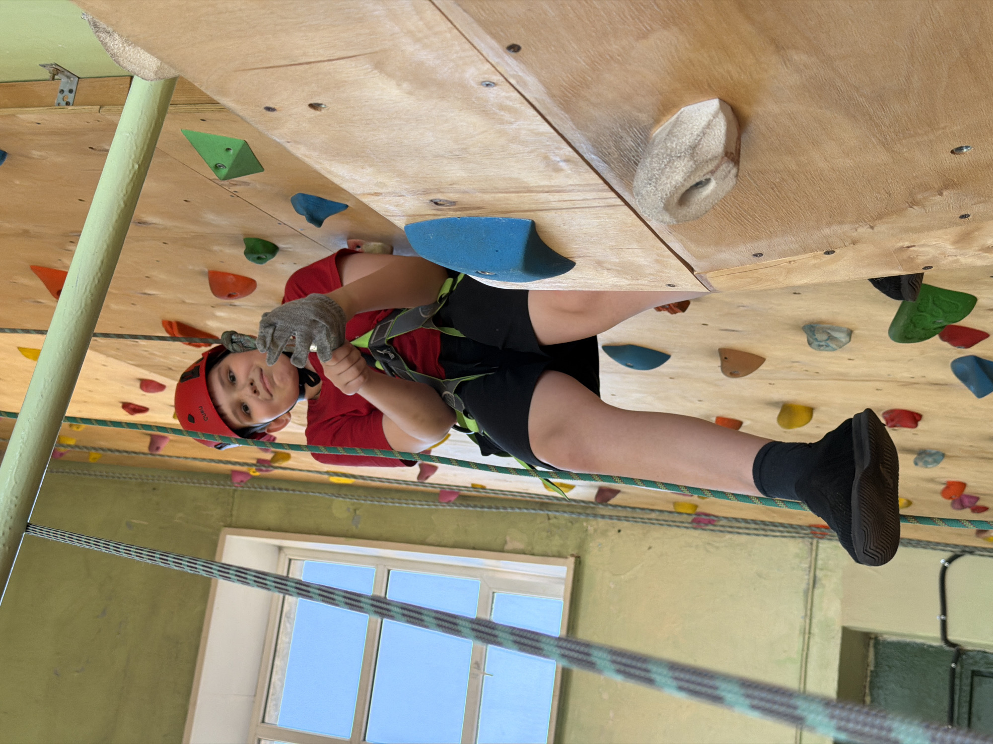 A child wearing a red climbing helmet, a red shirt, black shorts, and gloves, is rock climbing on an indoor climbing wall. The child is secured with a harness and rope. The climbing wall is made of wood and has various colored climbing holds. A window is visible in the background. The image appears to be rotated counter-clockwise. The location in Izhevsk, Russia is not known.
