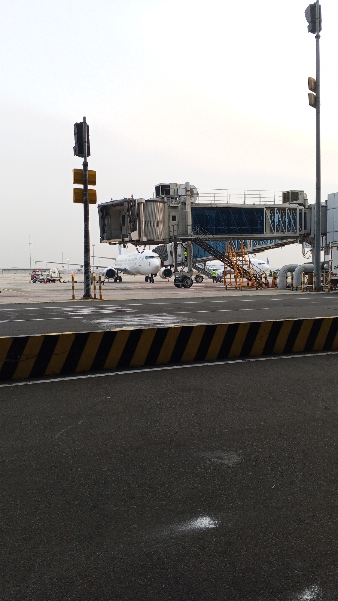 An airport scene is depicted from an eye-level perspective, looking across a tarmac towards a jet bridge connected to an airplane. The sky is overcast and bright.
In the foreground, a black asphalt surface is visible, marked with white lines and a section of yellow and black striped barriers.
Dominating the middle ground are two jet airplanes, a jet bridge, and airport service equipment. The jet bridge, a metallic structure with a blue-tinted glass enclosure, extends from the terminal building to the left of the frame and connects to the fuselage of the airplane on the left. The airplane is white with a red logo on its tail. A second white airplane is partially visible to the right of the first. Various service vehicles and equipment are positioned around the airplanes. A tall traffic signal with black and yellow lights stands to the left of the jet bridge.
In the background, more airport infrastructure is visible, including another airplane, airport buildings, and the hazy horizon. The overall environment suggests an active airport setting. There are no discernible individuals in the image performing specific actions. The scene is static and appears to be taken during daytime. The location is identified as Rawa Burung, Indonesia.