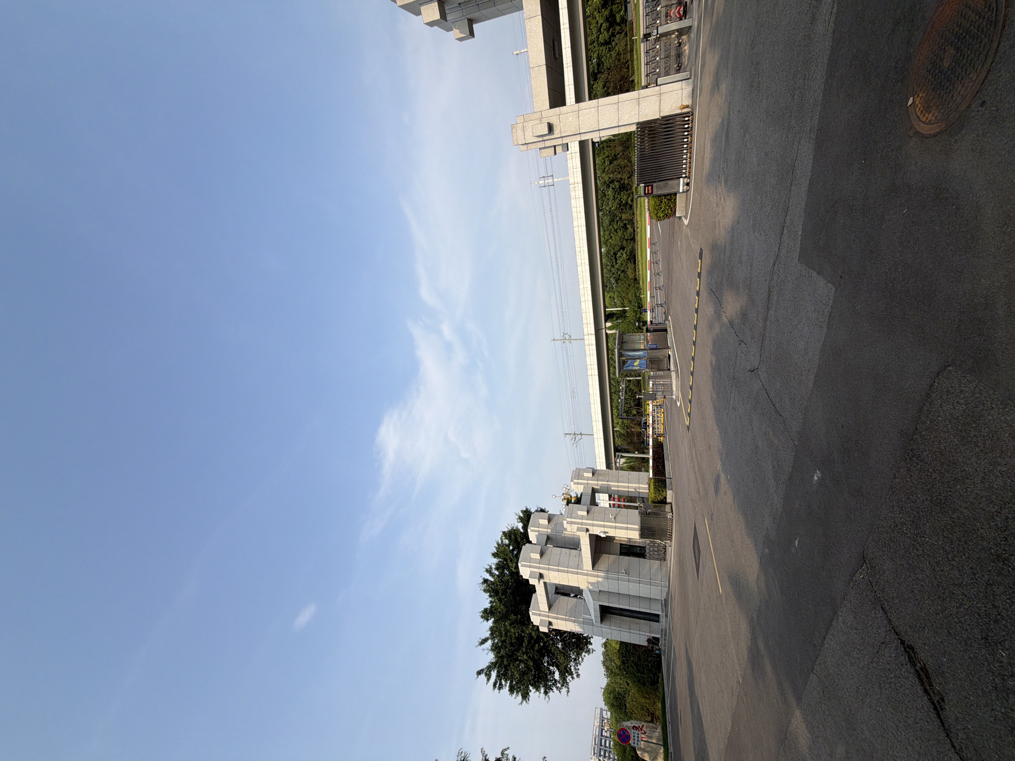 The image depicts the entrance to a modern facility in Jiangjin, China, under a clear blue sky with sparse clouds. The architecture is characterized by geometric, angular structures made of light-colored stone, resembling a gateway. A large, bushy tree stands to the left of the main structure. In the background, a railway bridge spans across the scene, with train tracks visible. The foreground is an asphalt road leading to the entrance, which features automated gates and bollards. A small guard booth is situated to the left of the entrance, with a traffic sign on a stone pillar to its left. The scene appears to be a controlled access point to a campus or industrial area, with no people clearly visible in the immediate vicinity. The environment suggests an urban or suburban setting.