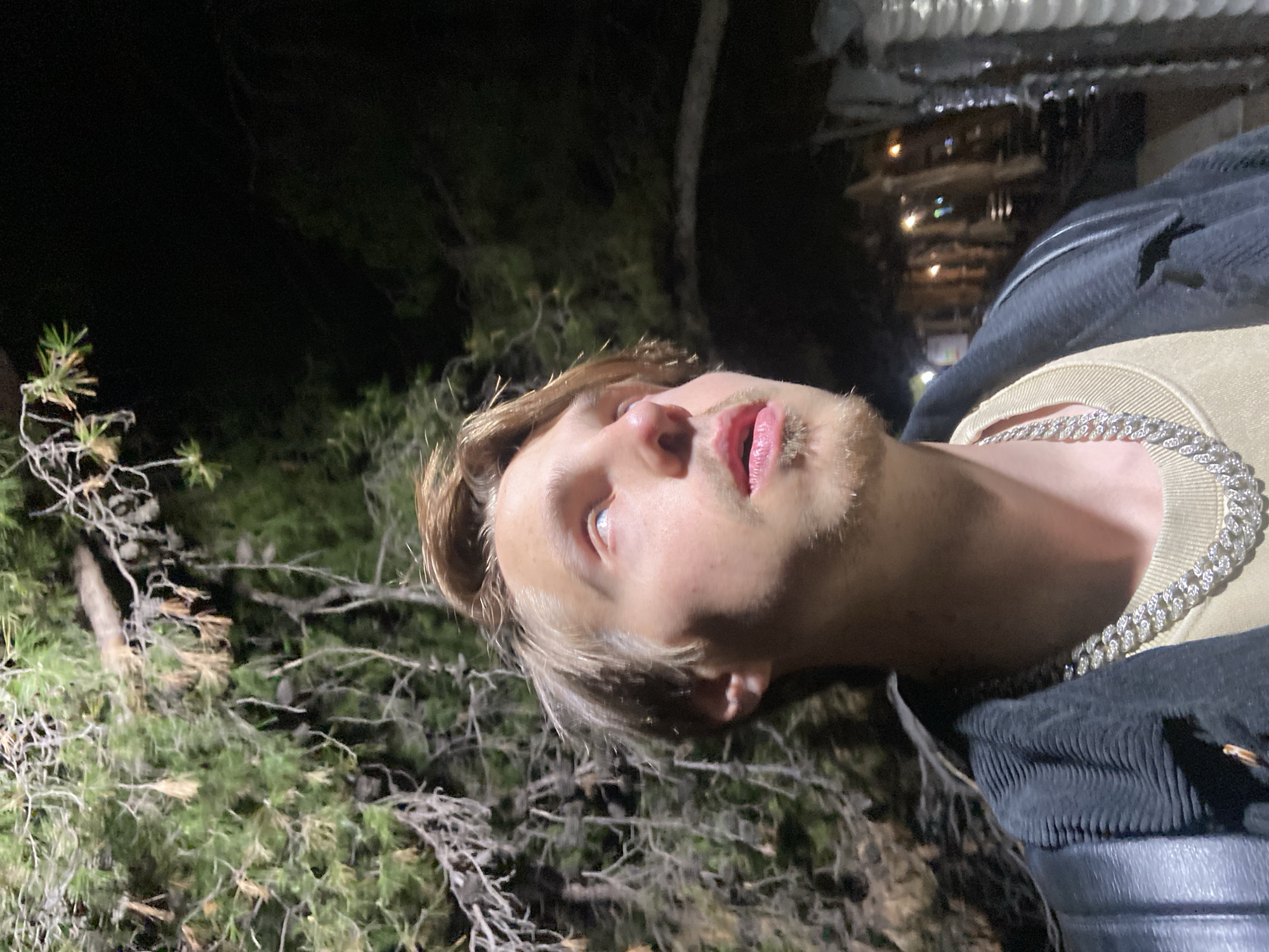 A nighttime outdoor scene shows a fair-skinned young adult male with blonde hair and stubble on his face. He is wearing a tan-colored shirt, a black corduroy jacket, and a silver chain necklace. The background is blurred, showing sparse vegetation and a building with lit windows. The building indicates this is an urban, potentially residential area. The angle of the image suggests the man might be lying down or tilting his head back. Although I lack sufficient information to pinpoint the exact location within Bar, Montenegro, the presence of vegetation and an urban structure could suggest it's at the city's edge or a park close to buildings.