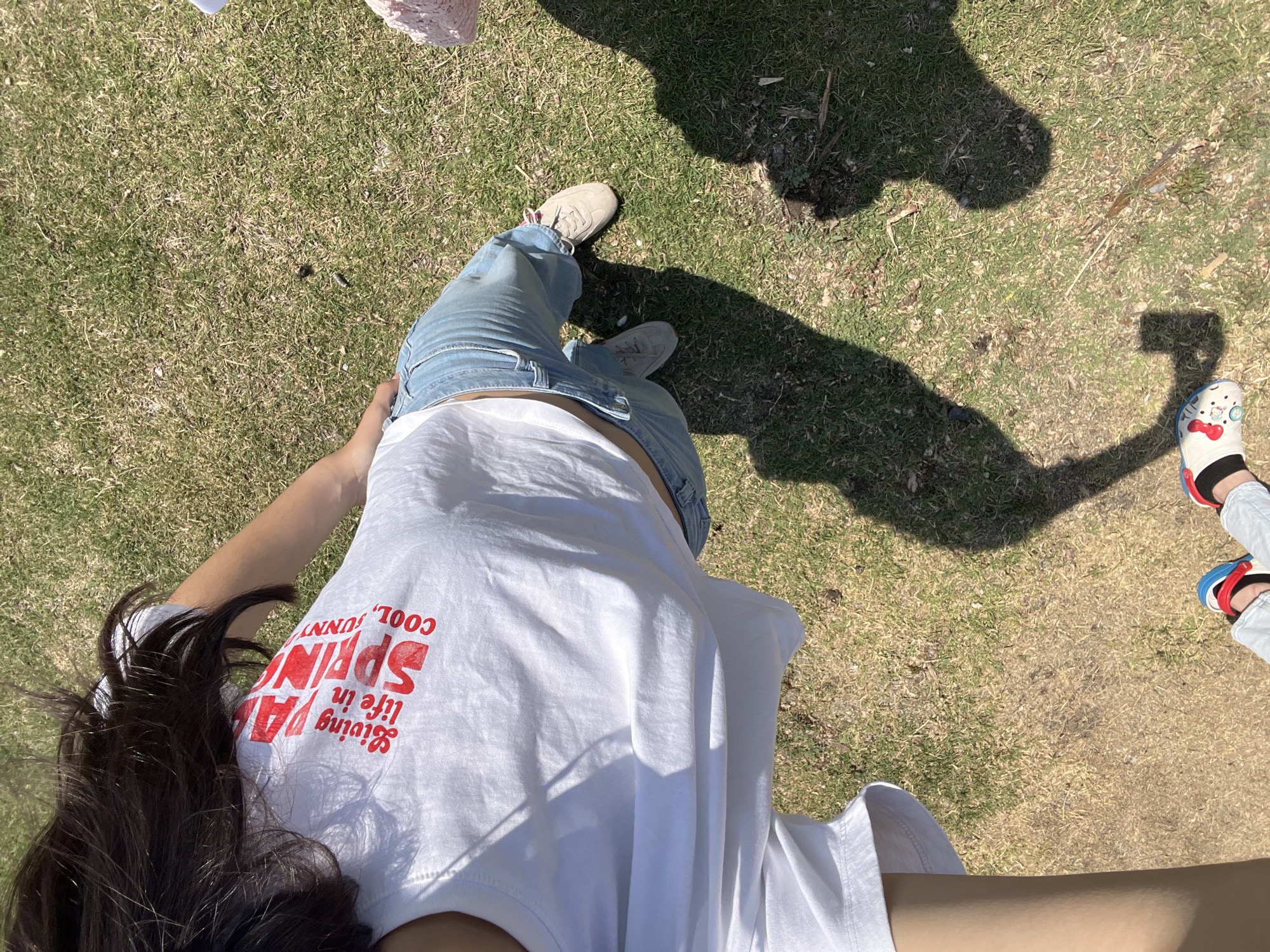 The image shows a first-person perspective of a person standing on grass, presumably in El Oasis Valsequillo, Mexico, given the prompt. The person is wearing light blue jeans, a white t-shirt with red text, and beige shoes. The text on the t-shirt reads "Living Ya Life in SPRING! COOL, SUNNY". The person's hair is dark and visible on the lower left. There are shadows visible on the grass, including the shadow of the person taking the photo and at least one other person wearing novelty shoes, red, white, and blue, standing to the right. The grass appears to be short and dry in places.