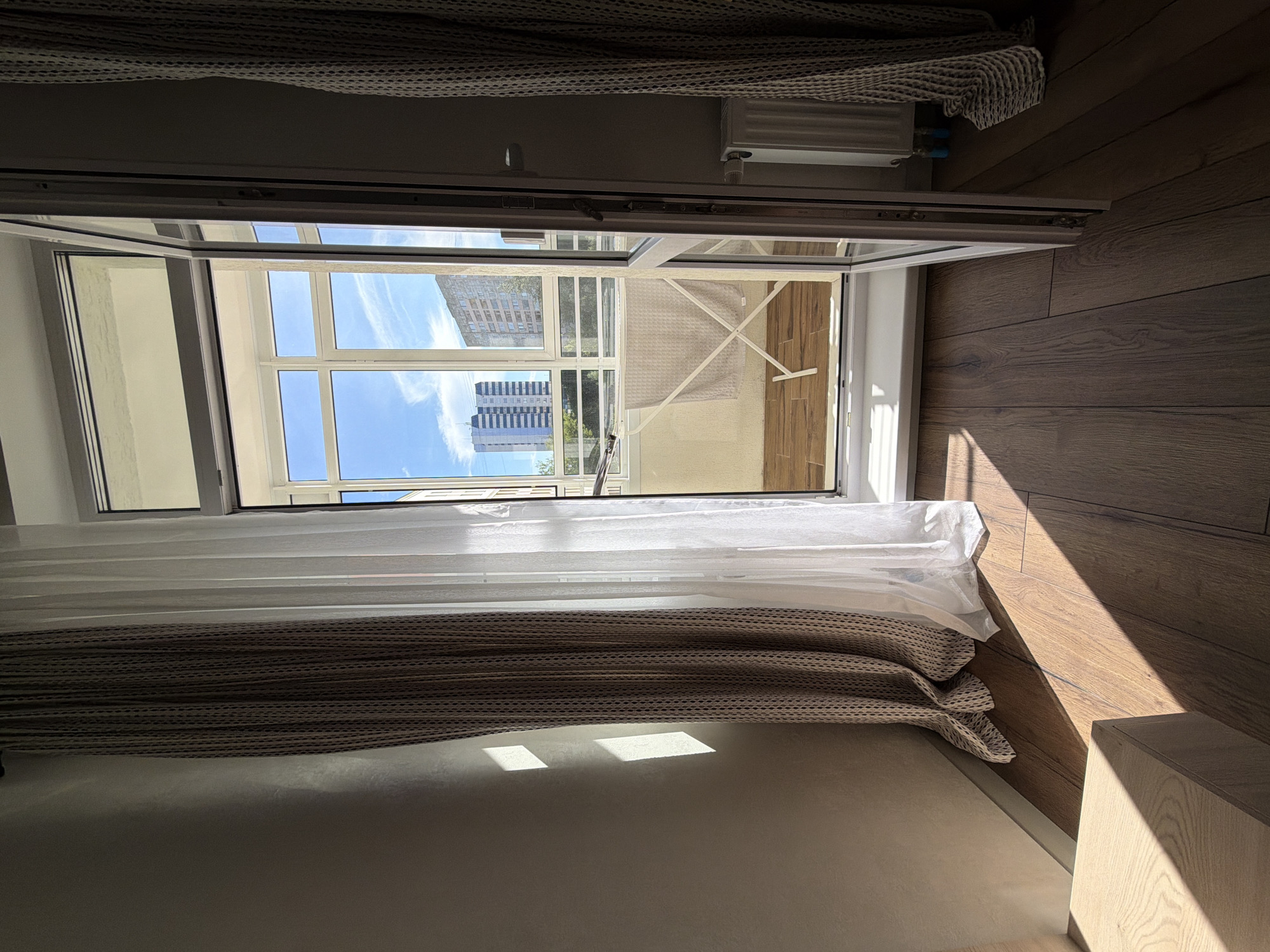 This image depicts an interior view of a room looking out onto a balcony and the cityscape of Perm, Russia. Sunlight streams through an open balcony door, casting strong shadows on the wooden floor and textured wall. A patterned curtain hangs on the left, and a sheer white curtain is draped near the balcony door. On the balcony, a drying rack with a light-colored towel is visible. Beyond the balcony, apartment buildings are seen under a bright blue sky with scattered clouds. A white radiator is present on the right side of the room. There are no people or animals visible in the image.