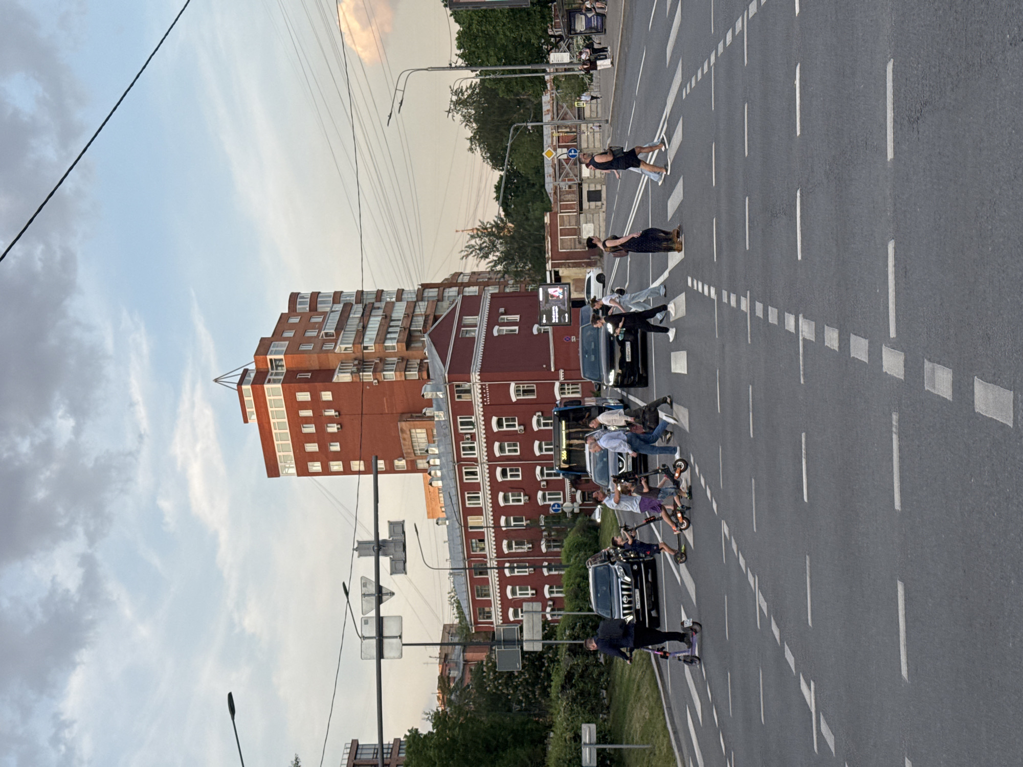 The image shows a street scene in Moscow, Russia. Pedestrians and scooter riders are crossing a wide road marked with white dashed lines for lanes and crosswalks. A brick building stands adjacent to the road, while streetlights and power lines are visible overhead. A black SUV is among the vehicles stopped to allow pedestrians to cross. The sky suggests either early morning or late afternoon light.