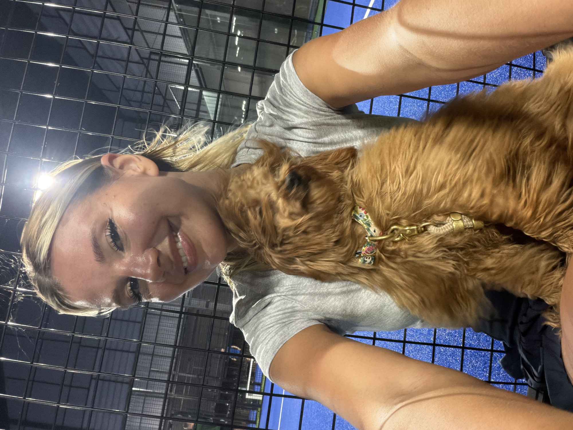 In Thep Krasatti, Thailand, a young woman with blonde hair pulled back in a ponytail takes a selfie with a small, fluffy brown dog. She is wearing a grey t-shirt and appears to be at a sports facility, as indicated by the blue court surface and black fencing in the background. The dog, wearing a decorative collar, is nestled in her arms, and its head is turned away from the camera. The lighting suggests it may be evening or night.