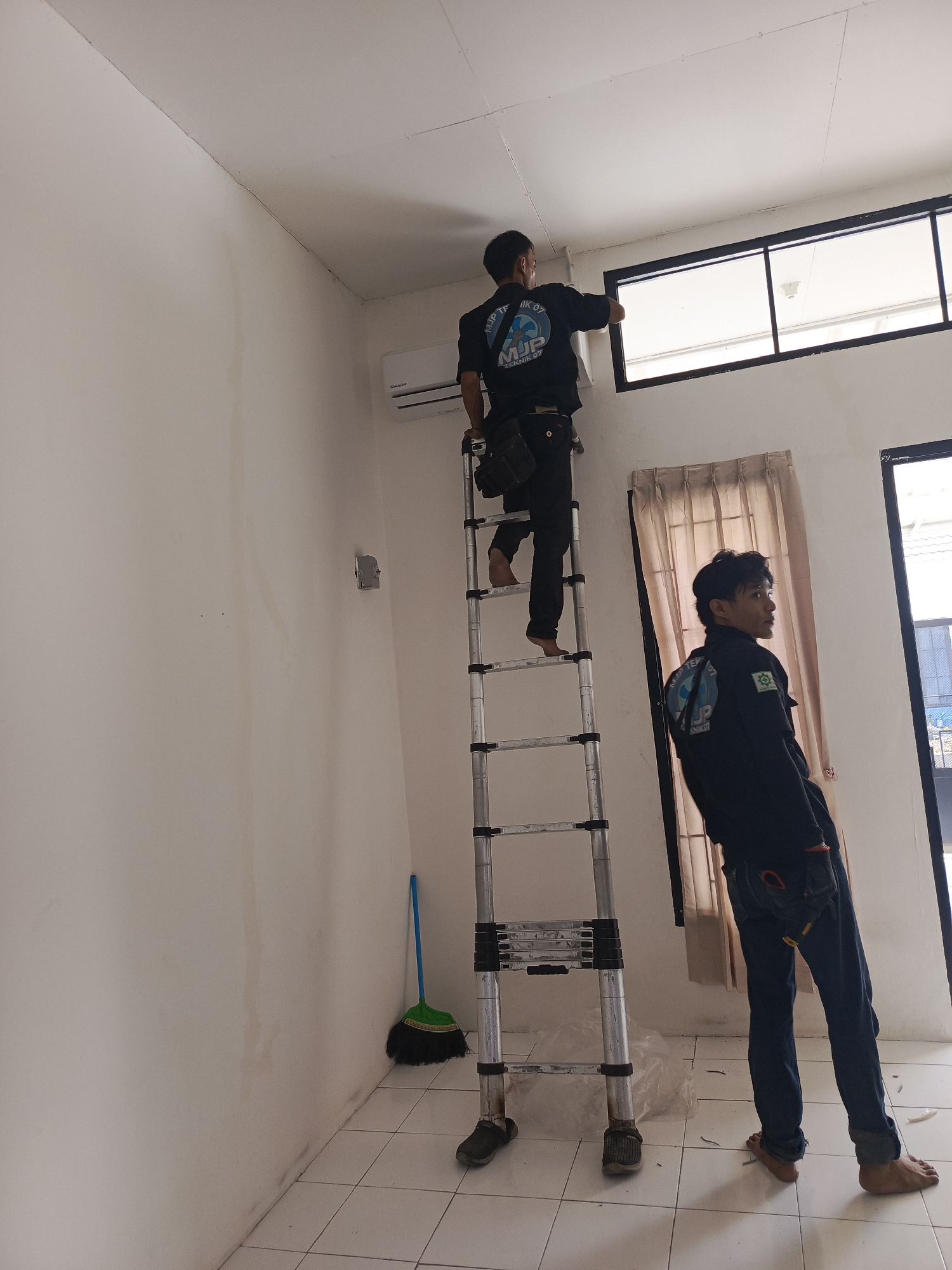 Two men in dark blue work uniforms are in a room with white walls and tiled floors. One man is standing on a tall, silver, extendable ladder, reaching up towards a window. He is wearing a tool belt. The other man is standing on the floor, looking to his right. He also has a tool belt on. A broom with a green handle and black bristles is leaning against the wall. A white air conditioning unit is mounted high on the wall. The room appears to be under renovation or construction, with some plastic sheeting visible on the floor. The scene suggests that the men are performing maintenance or installation work.