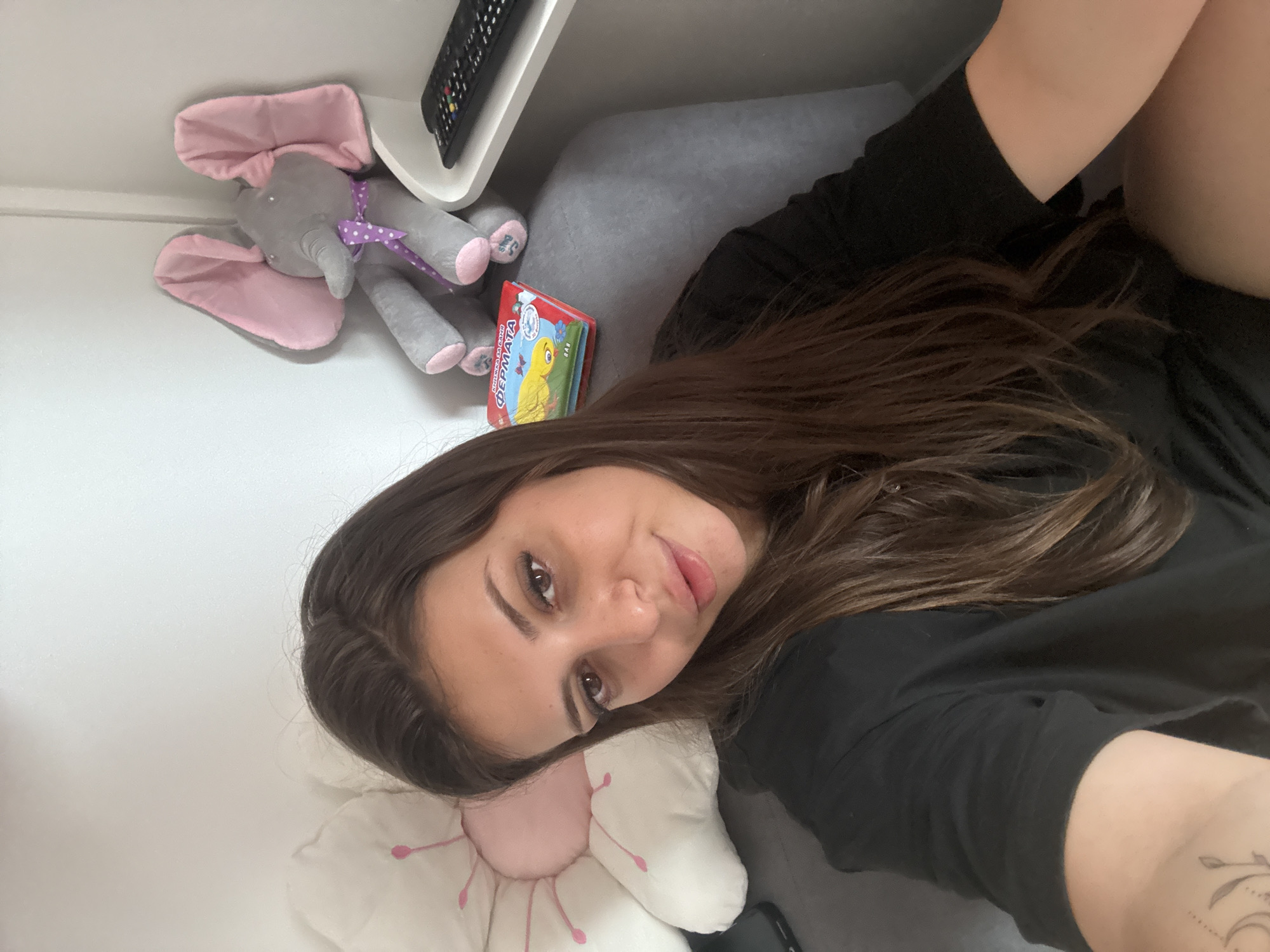 A young woman with long brown hair and dark eyes is sitting in a gray armchair. She is wearing a black t-shirt. To her left, there is a large, white, flower-shaped pillow. To her right, there is a stuffed gray elephant with pink ears and a purple polka dot ribbon around its neck, a small book, and a black remote control on a white shelf. The background appears to be a light-colored wall. The scene is indoors, likely in a home environment. No specific details about Plovdiv, Bulgaria, are visible in the image.
