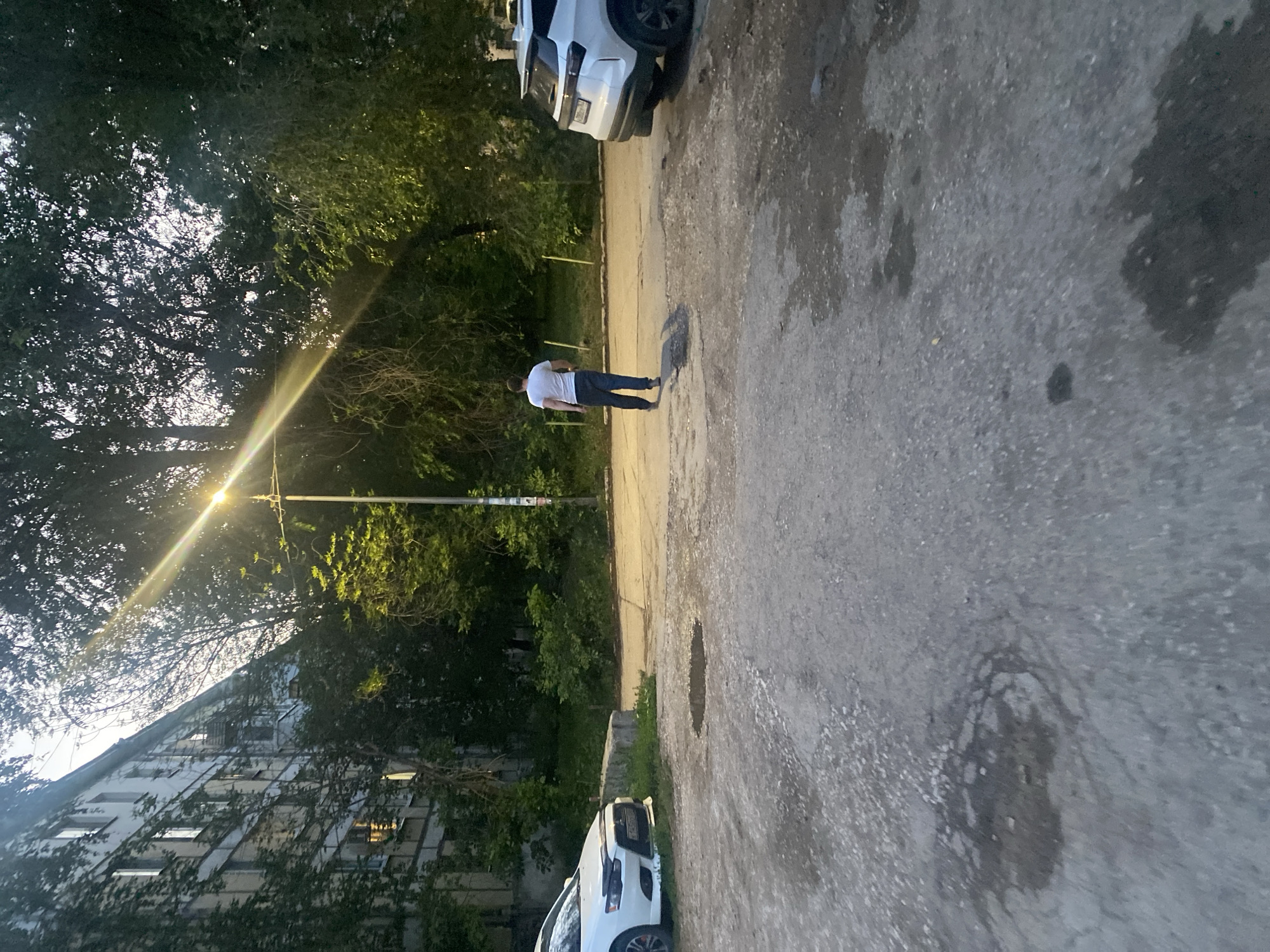 A medium shot shows a man walking away from the camera on a paved area in front of an apartment building. The man is wearing a white t-shirt and dark pants. The apartment building is to the left of the frame and has a light-colored facade with many windows. Trees with green foliage are visible behind the man and to the right of the frame, partially obscuring a street lamp with a bright light. A white car is parked to the left, and another white car is parked to the right, with its rear visible. The ground in the foreground is rough asphalt with several puddles. The scene appears to be in a residential area of Samara, Russia.