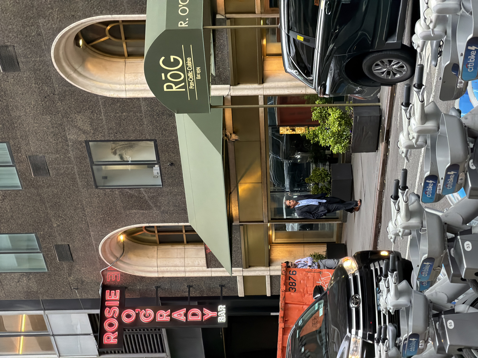 This urban street scene captures the entrance of a building featuring two distinct establishments, likely in New York City. The main subject is a modern building with a granite-like facade and arched windows on upper floors. A man in a dark suit and light blue shirt stands on the sidewalk near the building's entrance, looking towards the viewer.
Above the entrance, a dark green awning features the gold text "RŌG Pan-Celtic Cuisine Est 1974" and "R. O'C" (partially visible). To the left of the building's facade, a bright red neon sign vertically displays "ROSIE O'GRADY BAR". On the street level, a black car is parked, and a row of rental bicycles, identified as "Citi Bike" with some displaying numbers "53" and "55", line the sidewalk. Further down the street, an orange dumpster with the number "3876" is visible.
The lighting suggests it is daytime, possibly overcast or late afternoon, as indicated by the ambient light, yet the neon sign for Rosie O'Grady Bar is illuminated. The overall atmosphere is that of a bustling city street, with no specific activity beyond the man standing and parked vehicles.