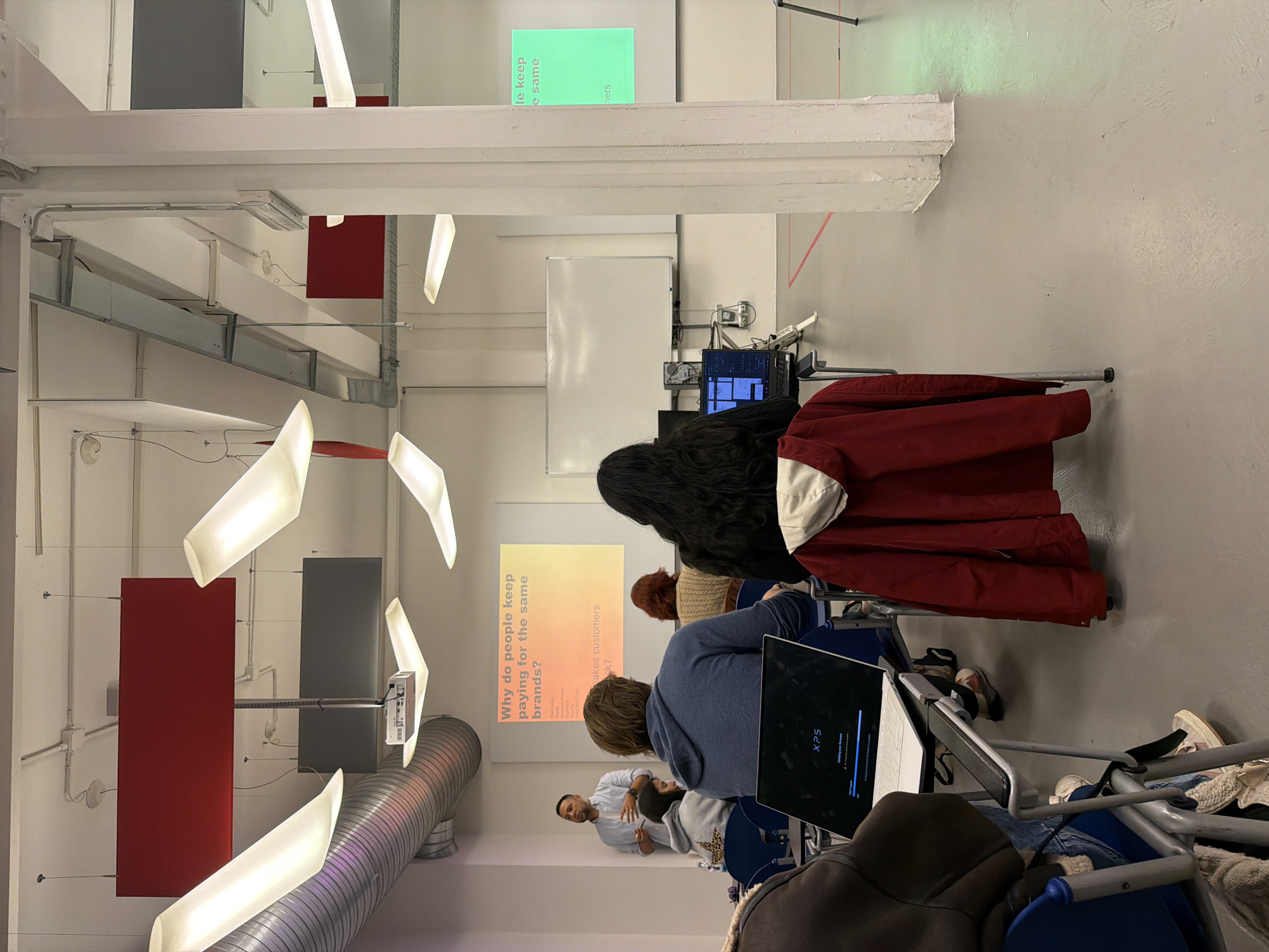 A classroom scene in Milan, Italy. A man is presenting information on a screen with the heading "Why do people keep paying for the same brands?". Several students are seated, facing the screen. Laptops are on the desks of several students. The room has a modern, industrial design with exposed ductwork and unique lighting fixtures.