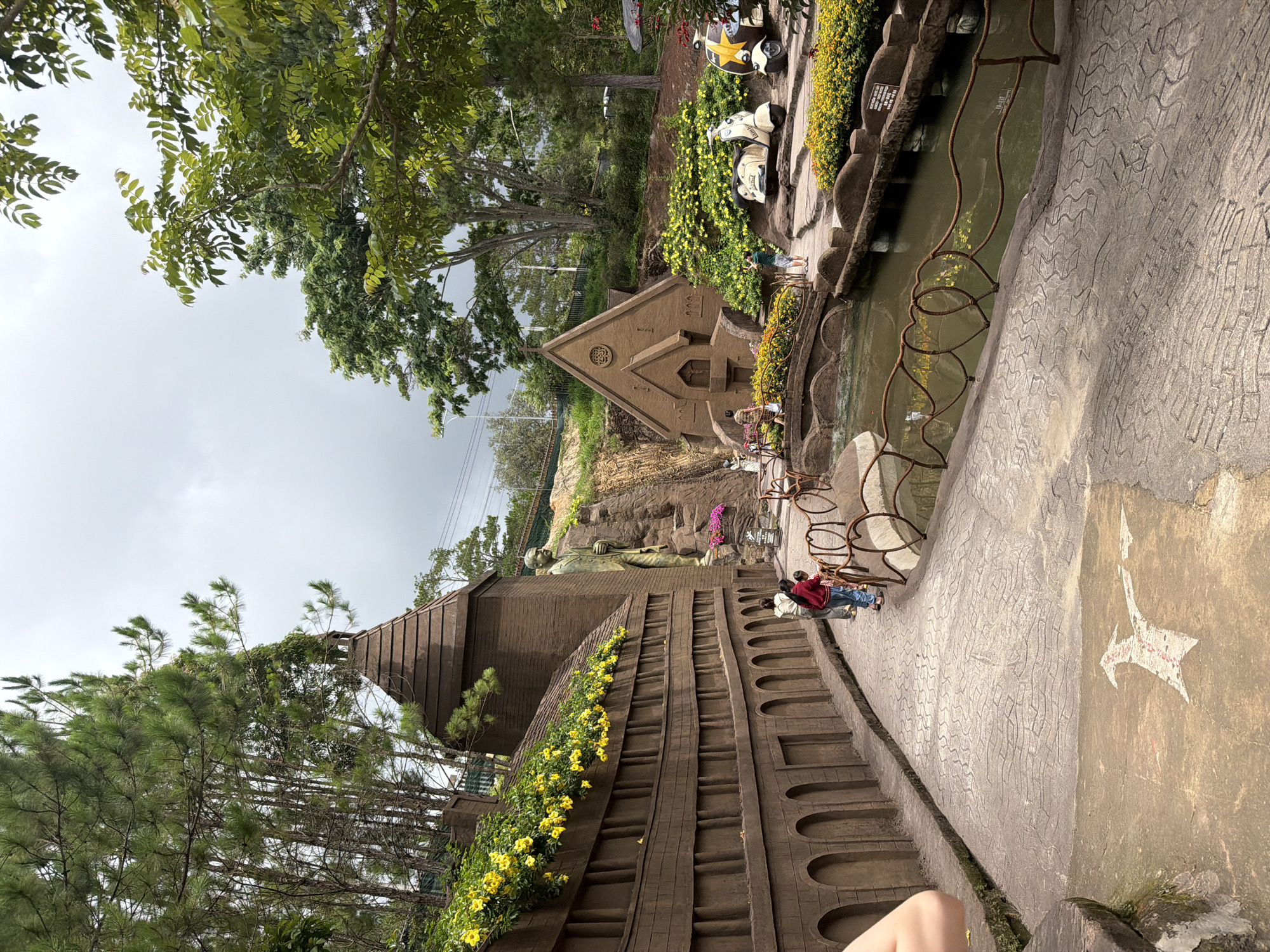 The image depicts a scene at the Crazy House in Da Lat, Vietnam, an architectural landmark characterized by its surreal and organic design. In the foreground, a textured concrete pathway curves to the right, marked with a white arrow pointing towards the upper right corner. To the left, a brown, brick-like wall with arched openings and decorative yellow flowers cascades down. A tall, bronze statue of a man holding a rifle stands to the right of this wall, partially obscured by a conical brown structure with a tiled roof.
Further into the scene, a small, brown, church-like building with a pointed roof and stained-glass windows is visible. A body of water, appearing as a pond or canal, runs through the middle of the image, spanned by a rustic bridge with decorative metal railings in the shape of hearts. Along the banks of the water are more flower beds filled with yellow blossoms. Two vintage-style scooters, one white and one with a yellow star, are parked near the church-like building.
Several people are present in the image. Three individuals, two adults and a child, are walking along the pathway near the large wall. Another person is seen taking a photograph near the bridge, and a few other figures are visible further in the background, suggesting a public park or attraction. The environment is lush and green, with various trees and foliage surrounding the structures, indicating an outdoor setting under an overcast sky.