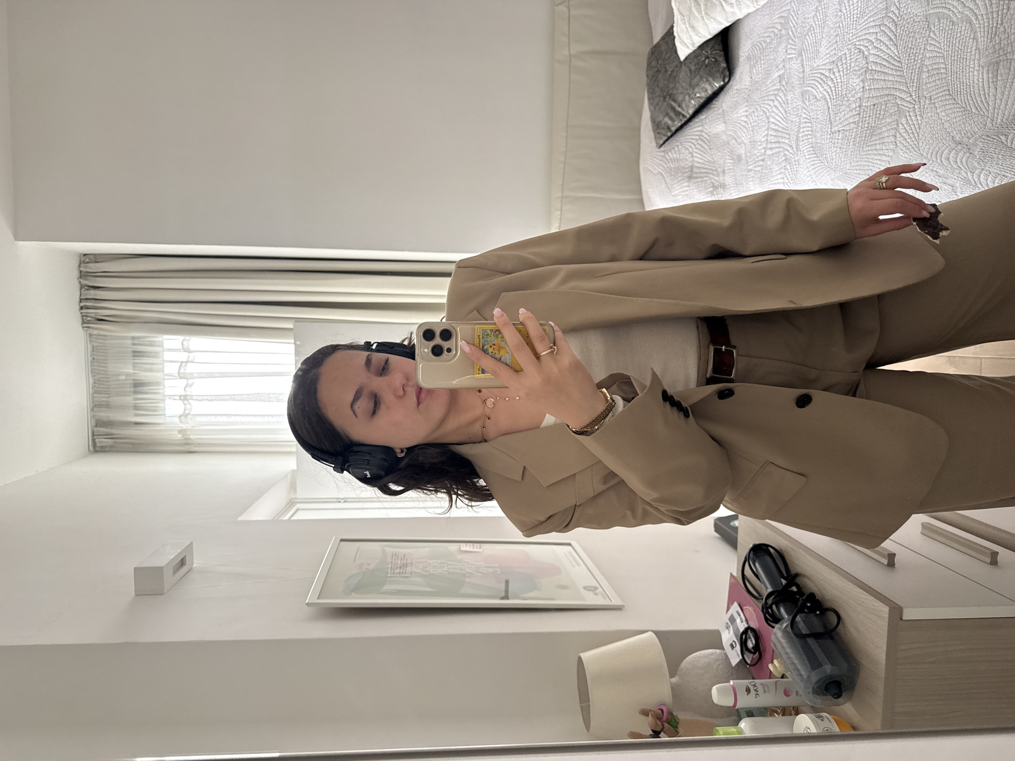 The image displays a young woman taking a mirror selfie in what appears to be a bedroom or dressing area. She is dressed in a light brown or tan pantsuit, featuring a structured blazer and matching trousers, worn over a white inner top. Black over-ear headphones are visible on her head, and her dark hair frames her face. Her eyes are closed or partially closed, suggesting a posed, relaxed, or contemplative expression.
She holds a gold-colored smartphone in her right hand, encased in a clear cover that prominently displays a yellow cartoon character, possibly Pikachu, resembling a Pokémon trading card. Her nails are neatly manicured with light pink and white tips, and she wears multiple gold rings on her fingers, including a distinctive knot-design ring. A gold bracelet and a watch adorn her left wrist, and a delicate gold necklace with small charms is visible around her neck. A brown belt with a silver buckle is cinching her trousers.
The background of the reflection shows a bright room, illuminated by natural light entering through a window fitted with light-colored blinds, indicating it is daytime. Behind the woman, a bed with white quilted bedding and a grey decorative pillow is neatly arranged. To her left, below the window, there's a white shelf holding a framed picture and a white rectangular object, possibly a speaker. To her right, a light wood-toned dresser or table is visible, cluttered with various personal care items including bottles, one clearly labeled "DOVE," a hairbrush, hair ties, and a black microphone-like object. A small lamp with a white shade also sits on the dresser. The room appears tidy and contemporary. There are no specific visual cues within the image itself to confirm the location as Milan, Italy.