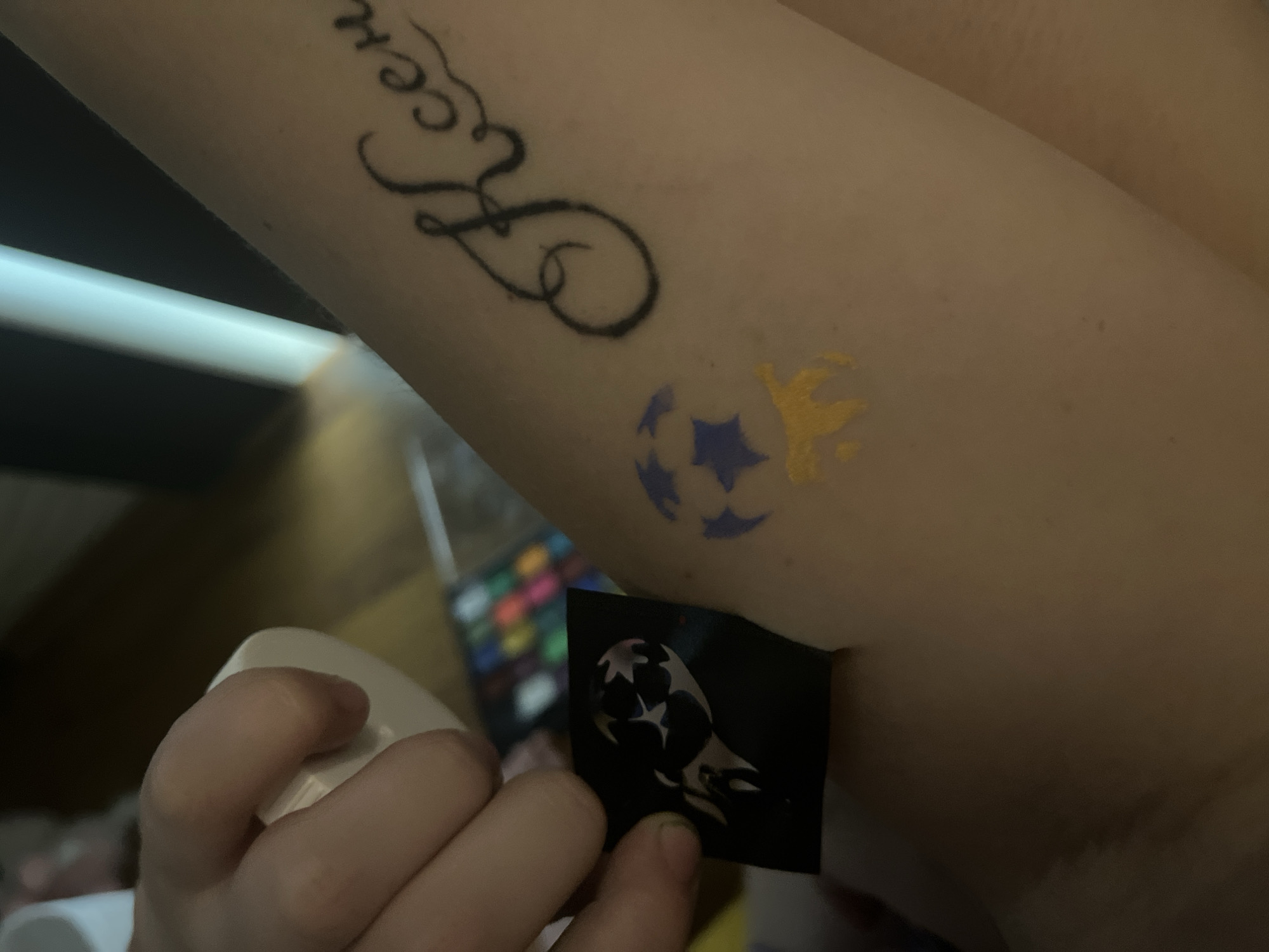A person's arm is visible in the foreground, with a temporary tattoo of a soccer ball with blue stars and yellow accents. A black stencil with a design that appears to be a stylized flame or wing is being held by a hand to the left of the arm. In the background, a black tattoo of the Cyrillic letters "Ксен" is visible on the arm. A blurred background suggests an indoor setting, possibly with a palette of colorful markers or paints. The image does not contain any visual information that would place it in Sochi, Russia.