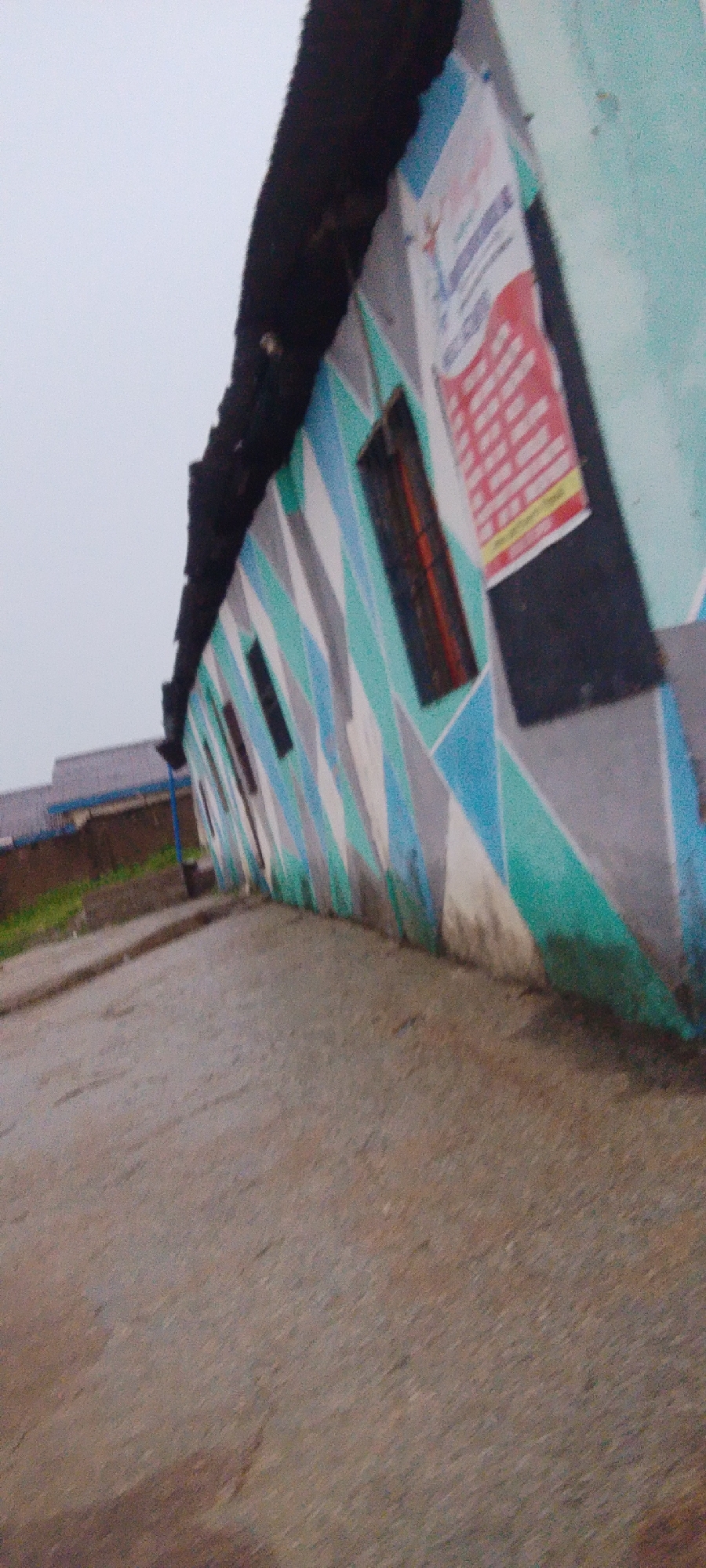 The image depicts the exterior of a building with a distinctive patterned facade. The building is angled, with a black, textured roofline dominating the upper portion of the frame. The walls are decorated with a geometric pattern of interlocking diamonds in shades of light blue, grey, and teal. A poster, predominantly red and white, is affixed to the wall on the right side of the image. Several dark, rectangular windows are visible along the building's side. In the background, a portion of another building with a grey roof and blue trim can be seen, along with some greenery. The foreground consists of a wet, grey paved surface, possibly a road or pathway, suggesting recent rainfall. The sky is overcast and a pale grey. There are no people or animals visible in the image. The scene appears to be in an outdoor urban or semi-urban environment.