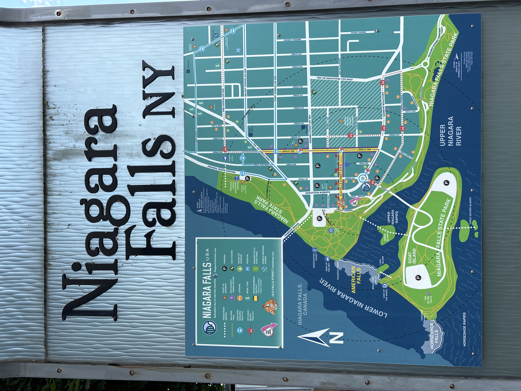 This media file is a photograph of a large, outdoor map sign for Niagara Falls, USA. The sign is vertically oriented and mounted on a textured, light blue wall, with a metal frame visible at the top and bottom. The map itself is predominantly green and blue, depicting the Niagara River and the surrounding park areas.
Prominently displayed on the left side of the sign are the words "Niagara Falls NY" in large, black, three-dimensional lettering. To the right of this text is a detailed map of the area, highlighting roads, points of interest, and geographical features like the American Falls, Horseshoe Falls, Goat Island, and Luna Island. A legend at the bottom left of the map explains various icons for services and attractions such as parking, restaurants, lodging, and transportation.
The map also includes text labels for streets, parks, and water features. Several key locations are marked with icons and text, including the Niagara Falls Visitor Center, Niagara Falls State Park, and the Seneca Niagara Casino. The Upper Niagara River and Lower Niagara River are clearly delineated. A small section on the upper right of the map is labeled "Other Points of Interest (North)," and below it, a list of destinations is provided.
The overall scene suggests a tourist information or welcome sign, likely situated near an entrance to the park or a visitor area. The lighting appears to be natural daylight, and the weather seems clear. There are no people visible in the photograph, making the map and its informational content the sole subjects. The sign appears to be relatively new and well-maintained, despite some minor wear and dirt visible on the blue wall behind it.