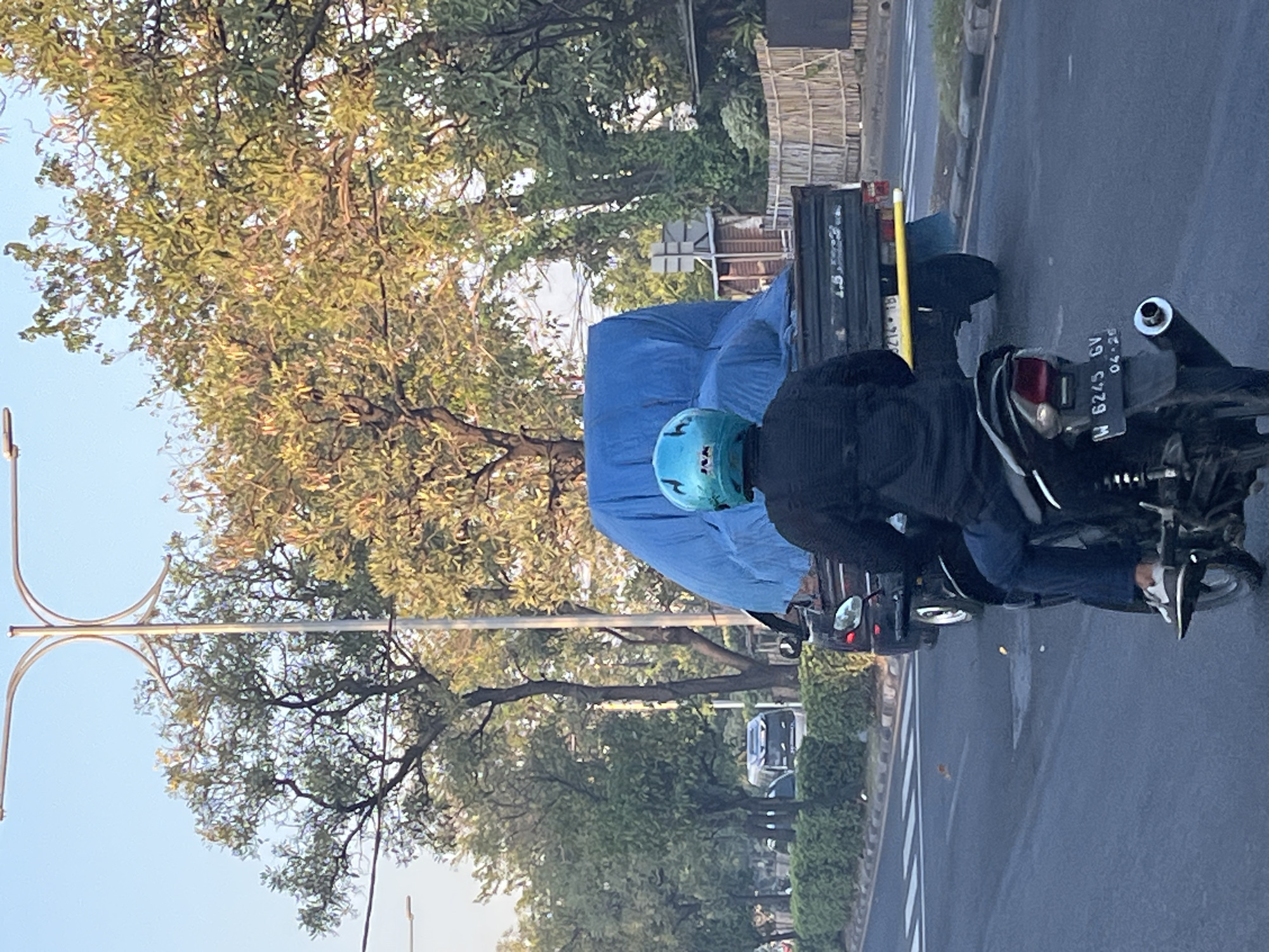 In the city of Kludan, Indonesia, on a street with a light blue sky and leafy green trees, a motorcycle rider wearing a blue helmet with "J&K" branding is facing away from the camera. The rider, dressed in a dark jacket and blue jeans, sits on a black motorcycle with a visible license plate that reads "W 6245 GV 04-26". In front of the motorcycle, a black truck is partially visible, covered with a large blue tarp. The truck has a yellow bar extending from its rear and a license plate that appears to be "214 1B". To the left of the truck, a small bus or van is seen on the road. The street has a white crosswalk and is lined with manicured green bushes on the side. A wooden fence can be seen in the background behind the truck.