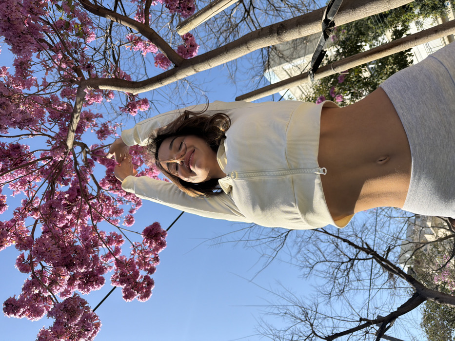An adult female is depicted stretching outdoors in an urban setting in Tel-Aviv, Israel. The subject, viewed from a low angle, wears a light cream ribbed cropped long-sleeve jacket and light gray athletic bottoms, revealing her midriff. Her eyes are closed, a subtle smile is present, and her arms are extended upwards with hands interlocked, suggesting a stretch. Above her, a tree with dense clusters of vibrant pink flowers blooms against a clear blue sky. To the left, a bare deciduous tree is visible. On the right, a supported tree trunk stands, with a light-colored multi-story building partially visible in the distant background. A thin black utility wire crosses the upper left of the frame. The scene is brightly lit by natural sunlight.