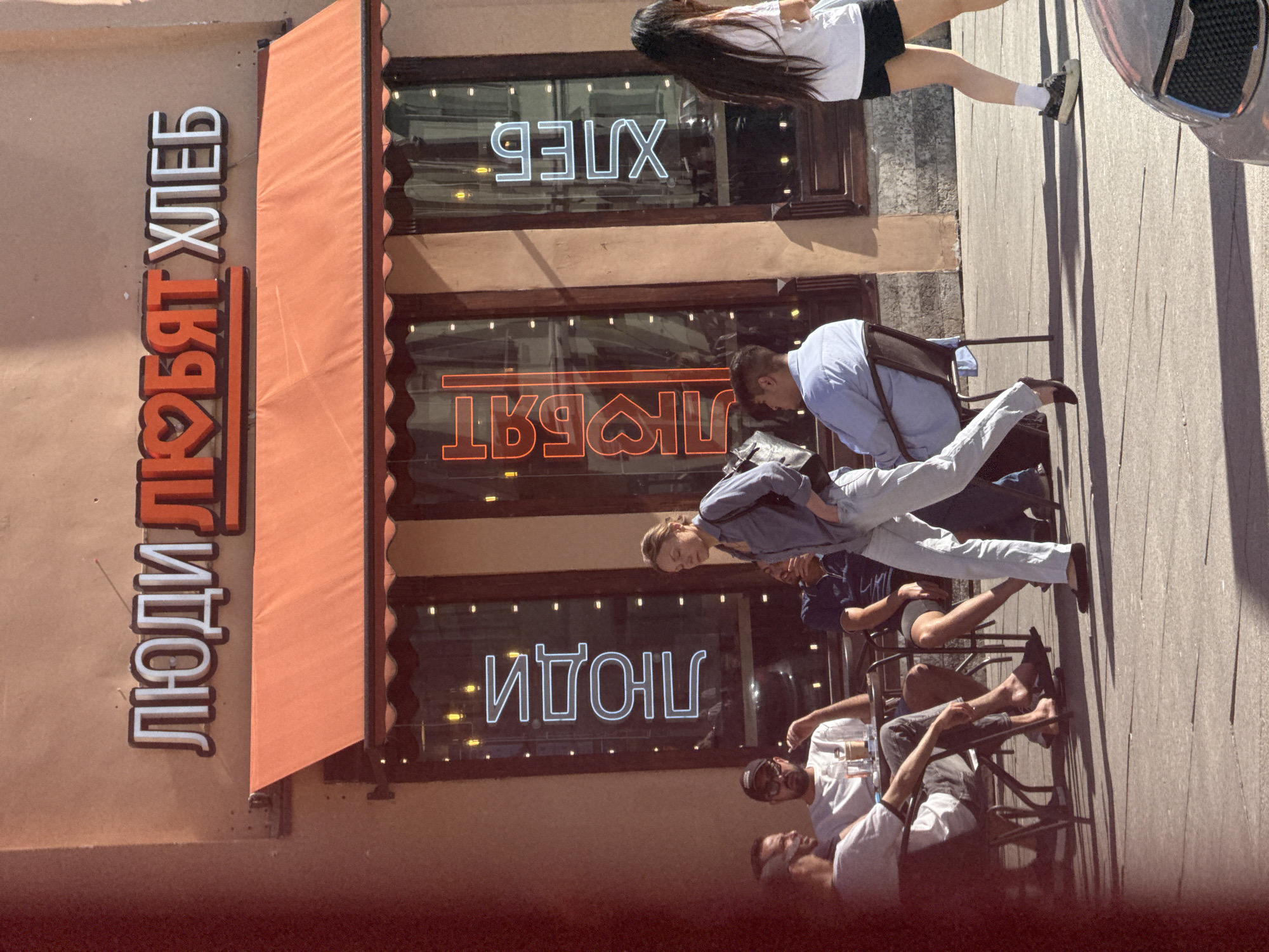 The image captures a street scene in Saint Petersburg, Russia, featuring a cafe with illuminated signage. Above an orange awning, the sign reads "ЛюДИ ЛЮБЯТ ХЛЕБ" (People love bread), with the word "ЛЮБЯТ" (love) stylized to include a heart symbol. Below the awning, vertical neon signs on the windows read "ЛЮДИ" (people), "ЛЮБЯТ" (love), and "ХЛЕБ" (bread) from left to right.
Several individuals are present. In the foreground to the left, a group of four men are seated at tables outside the cafe, engaged in conversation. One man wears a black baseball cap. A woman in a blue shirt and light-colored pants walks past the cafe entrance. To the right, a woman with long dark hair, wearing a white t-shirt and black shorts, is seen walking away from the camera. The cafe appears to be situated on a sidewalk with a paved street in the foreground. Shadows suggest it is daytime.