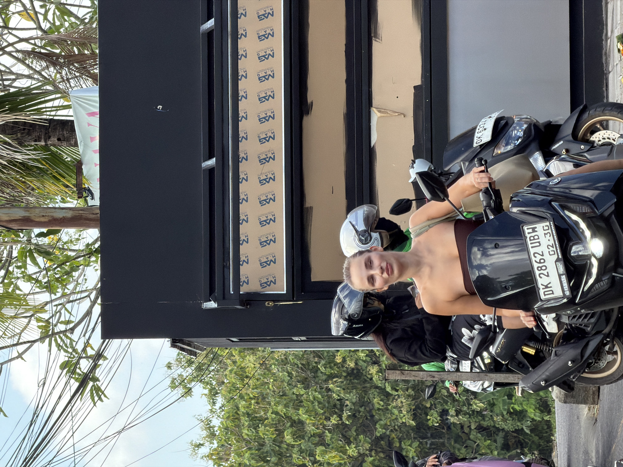 In North Kuta, Indonesia, a woman with blonde hair pulled back, wearing a brown strapless top and a light-colored bag slung over her shoulder, is seated on a dark-colored scooter. She is looking towards the camera and holding the handlebars. To her left, another person wearing a black hoodie and a black helmet with a clear visor is also on a scooter, facing forward. A third scooter, partially visible to the left, has a person wearing a purple outfit. Behind them, a building with a dark facade features a row of aluminum framing. The background is filled with lush green trees and overhead utility lines against a blue sky with scattered clouds. The scooters have license plates that read "DK 2862 UBW" and "DK 6334".