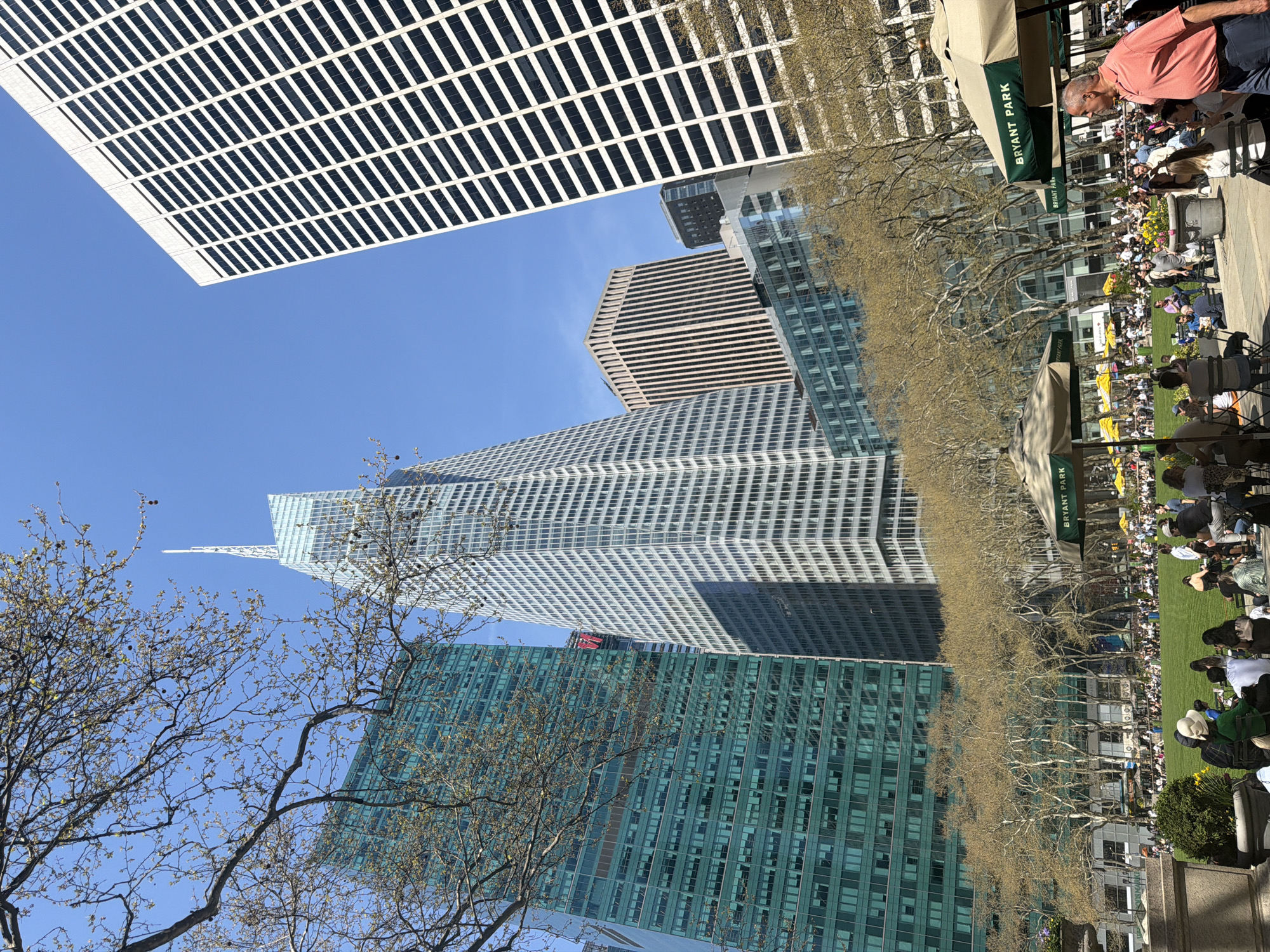 The photograph depicts a view from Bryant Park, New York City, United States of America. The foreground shows a crowd of people gathered in the park, possibly enjoying the outdoor space. The midground consists of trees lining the park with bare branches. The background features a collection of modern skyscrapers of varying designs and colors. The sky is blue and clear.