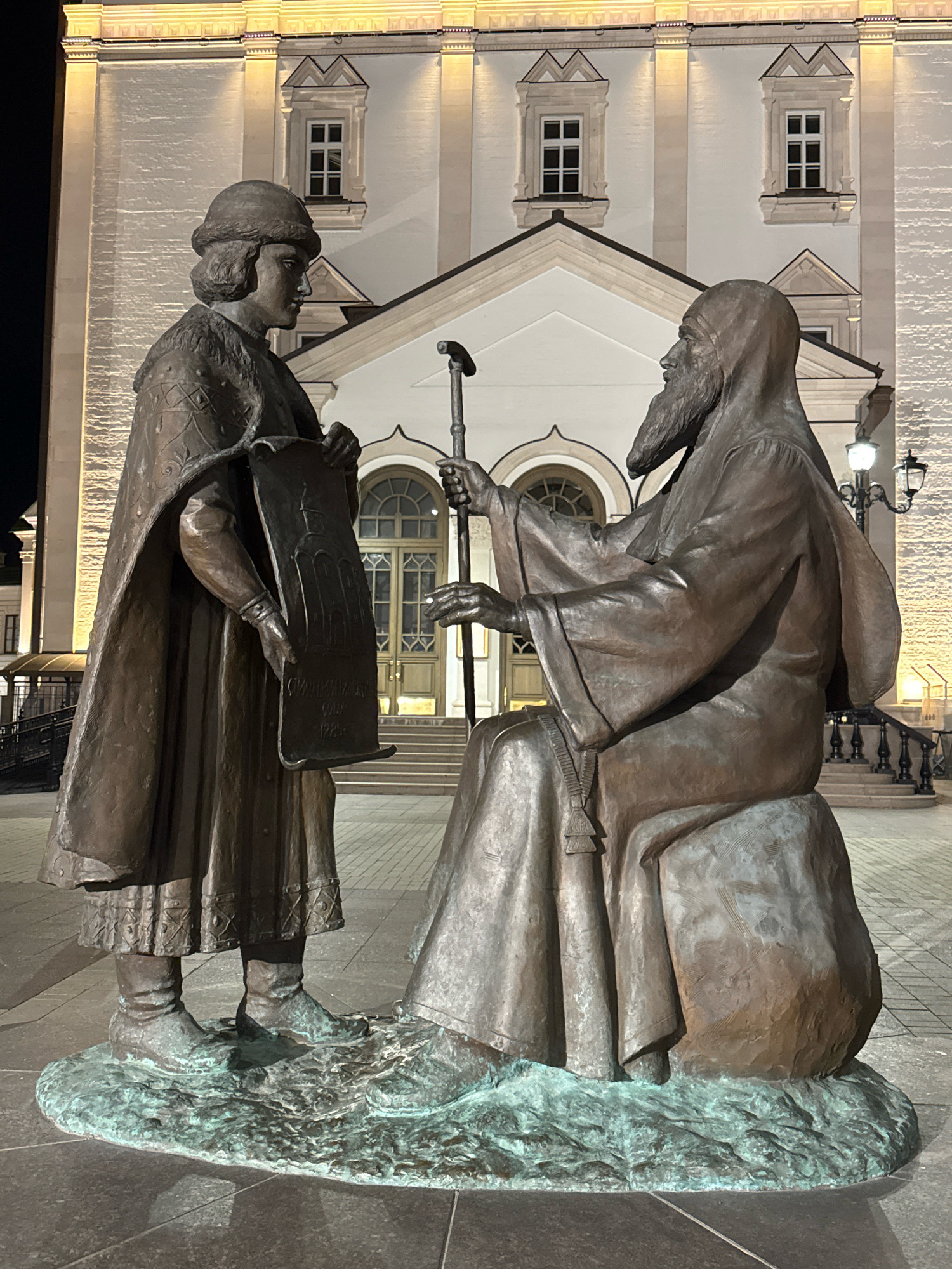 This is a medium shot of a bronze sculpture of two figures in front of a building at night. The sculpture depicts an older man with a beard, wearing a hooded robe, sitting on a rock and holding a walking stick. He is looking at a younger man standing in front of him, who is wearing a fur-lined cloak and a hat, and holding a scroll. The younger man is looking at the older man and appears to be presenting the scroll. The building behind them is lit and appears to be a historical or religious structure with arched windows and decorative elements. The ground in front of the sculpture is paved with stone tiles. This sculpture is located in the city of Tver, Russia.