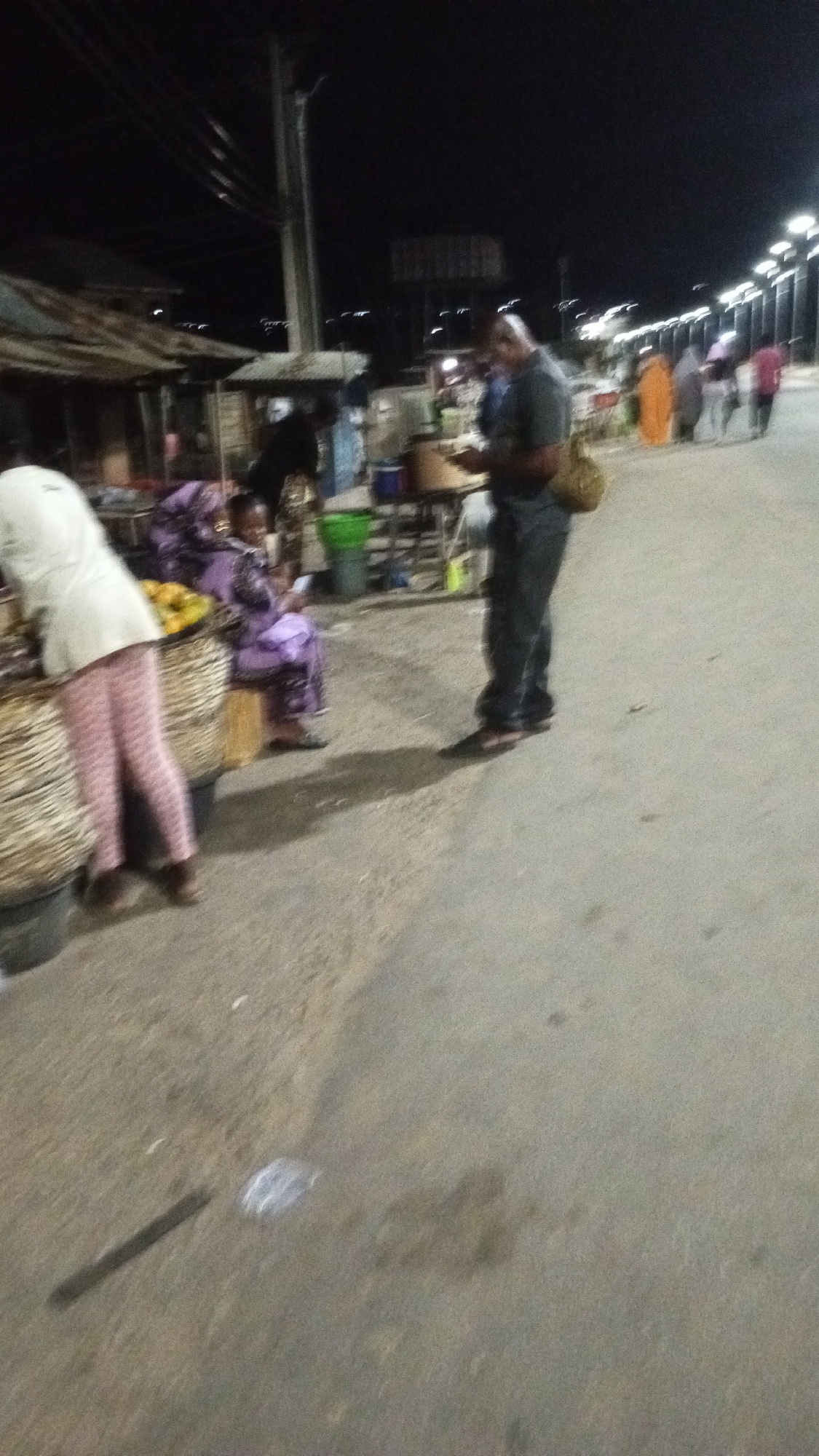The image captures a bustling street market scene at night in Adankolo, Nigeria. Multiple individuals are visible, engaged in various activities under the artificial glow of streetlights.
In the foreground left, a person wearing a white top and pink striped pants is bent over a market stall or large basket, possibly attending to goods. Next to them, a person in a purple patterned outfit is seated, looking down at a mobile phone. Further right, a man in a dark grey t-shirt and jeans, carrying a light brown bag over his shoulder, stands facing towards the market stalls. He is also looking down at an object in his hands, possibly a phone or a small item he has purchased.
The street is paved, and market stalls with various goods, including baskets of what appear to be yellow fruits or produce, line the left side. Green and blue buckets are also visible near the stalls. In the background right, several other people are walking along the road, some appearing to be moving away from the viewer. One figure is notably dressed in a bright orange or yellow garment. A series of streetlights illuminate the road, indicating it is nighttime. The overall scene suggests a vibrant, informal market or commercial area active after dark. No visible text is discernible in the image.