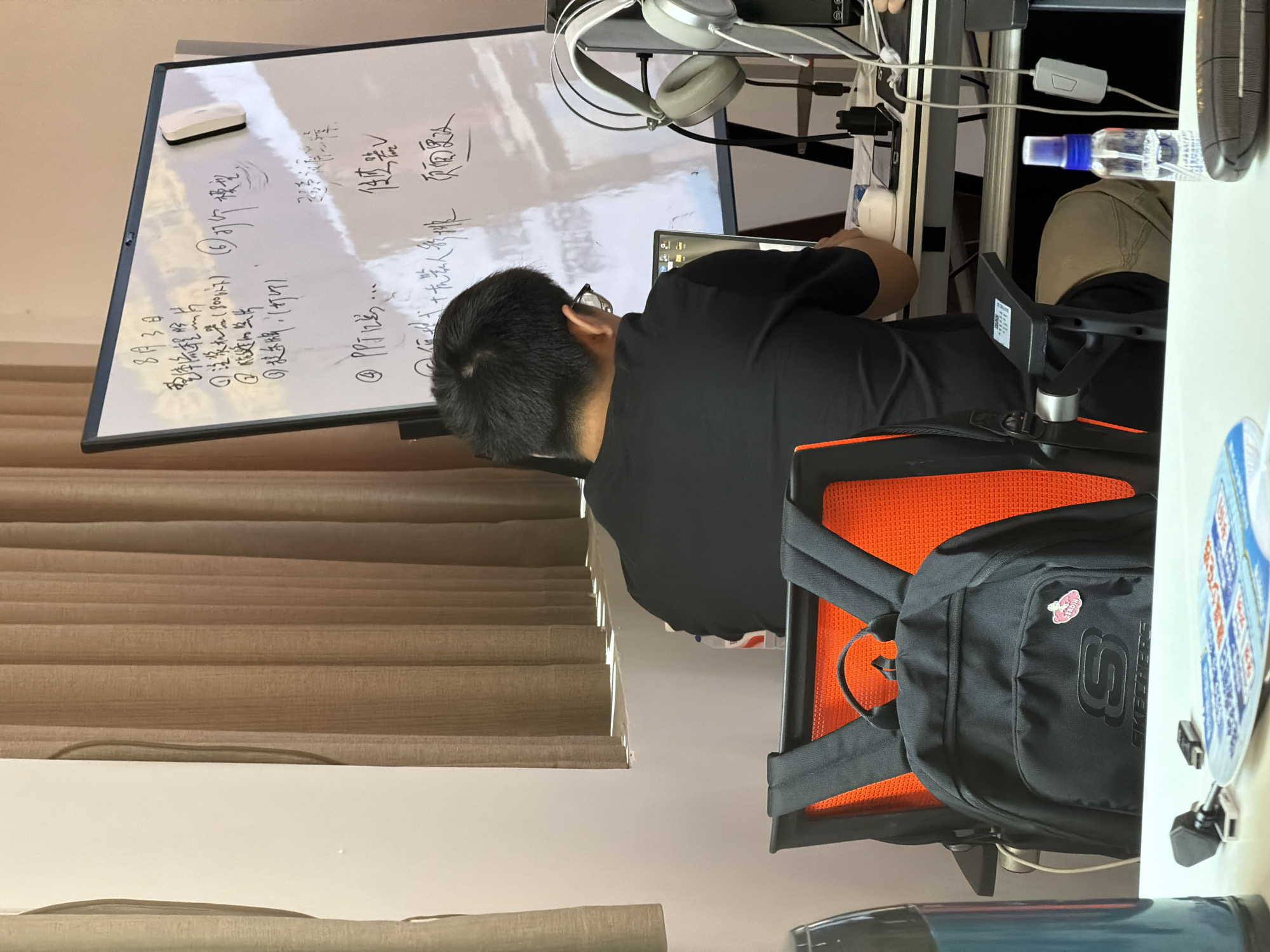 A man is sitting at a desk facing a laptop with a whiteboard behind him. The whiteboard has writing and drawings on it. The man is wearing a black t-shirt and glasses. He has a backpack on his chair. The environment appears to be an office or study. The writing on the whiteboard suggests it is a to-do list or meeting notes. A bottle of hand sanitizer is on the desk.